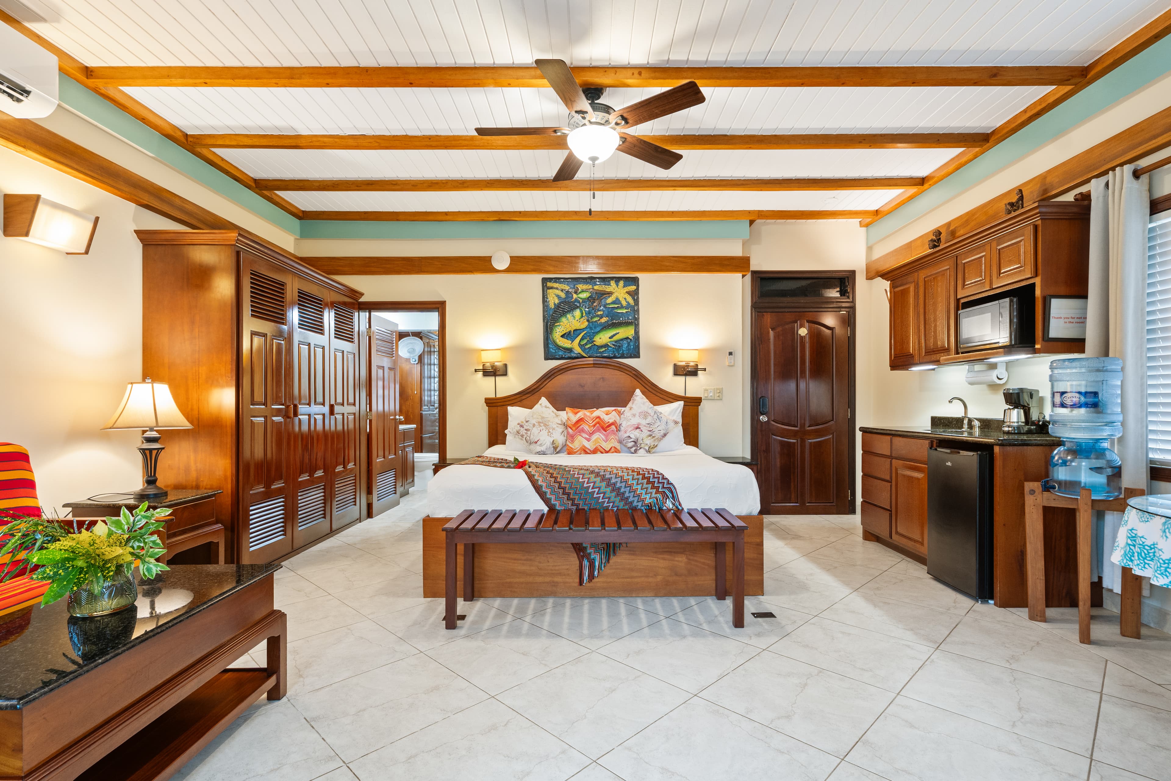 A third-floor guest room featuring a king-size bed with colorful accent pillows, wood-beamed ceilings, and a ceiling fan. To the right is a small kitchenette with a mini-fridge and microwave, and to the left is a large wooden wardrobe and a seating area.