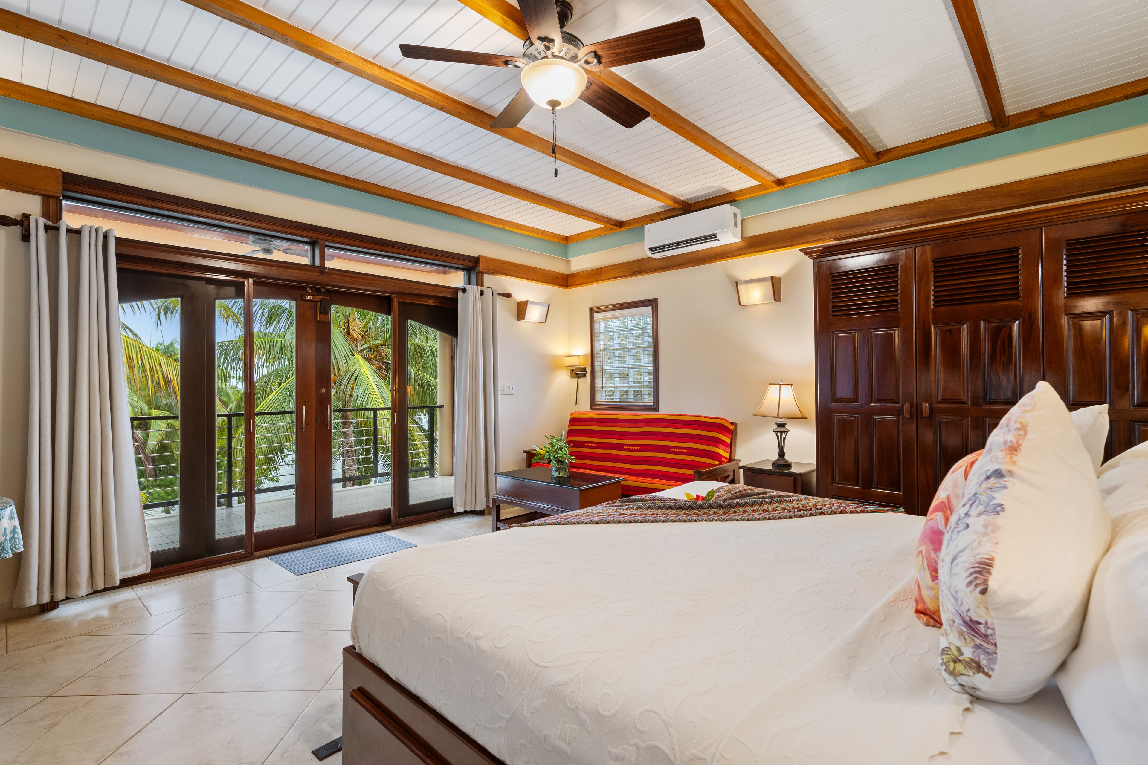 A king guest room featuring a private balcony with glass doors overlooking palm trees. The room includes a wood-beamed ceiling with a fan, a striped orange sofa, and large dark wood closets.