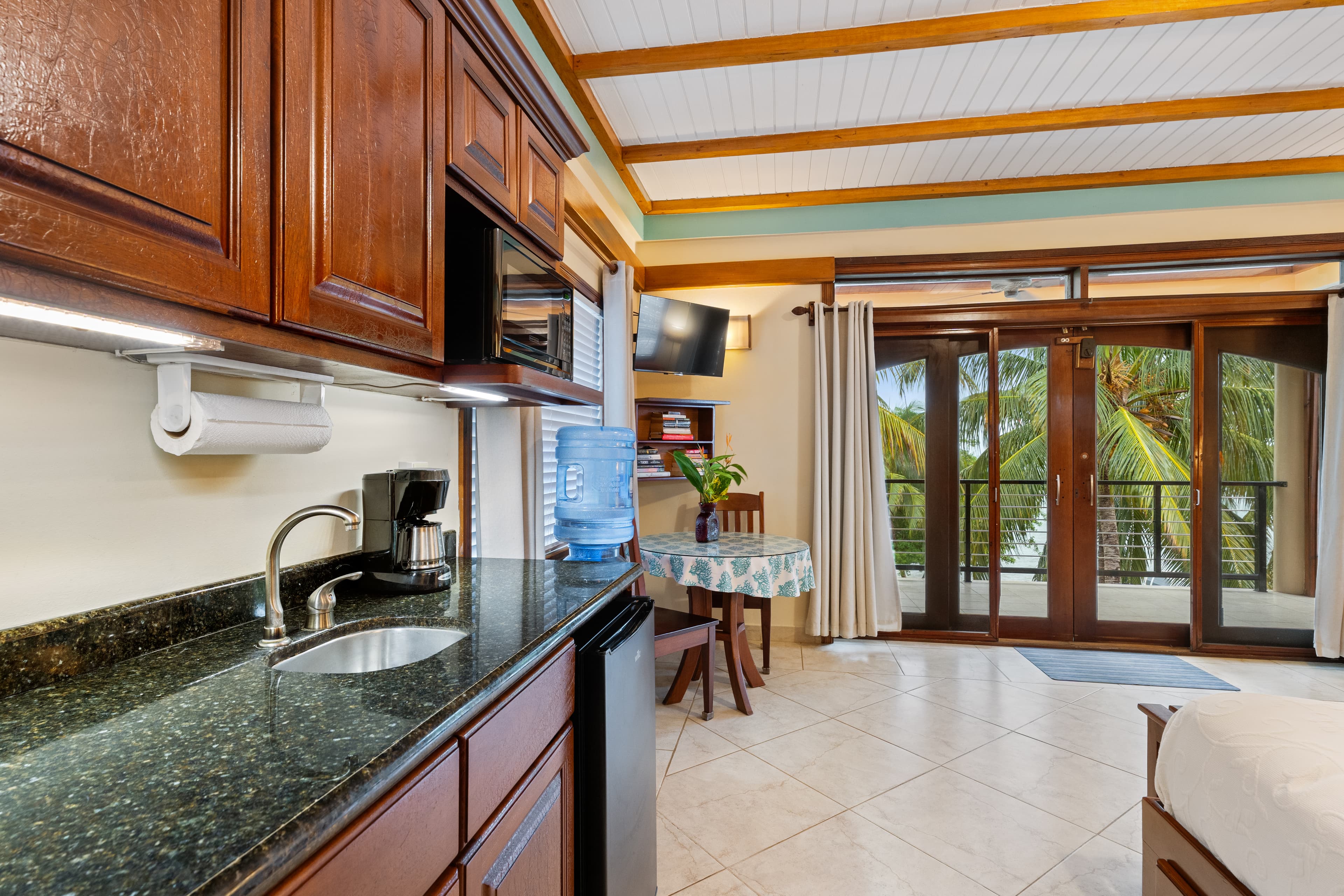 A kitchenette featuring dark wood cabinets, a black granite countertop with a small sink, and a coffee maker. In the background, a small dining table with a blue-patterned cloth sits near glass balcony doors that open to palm trees.