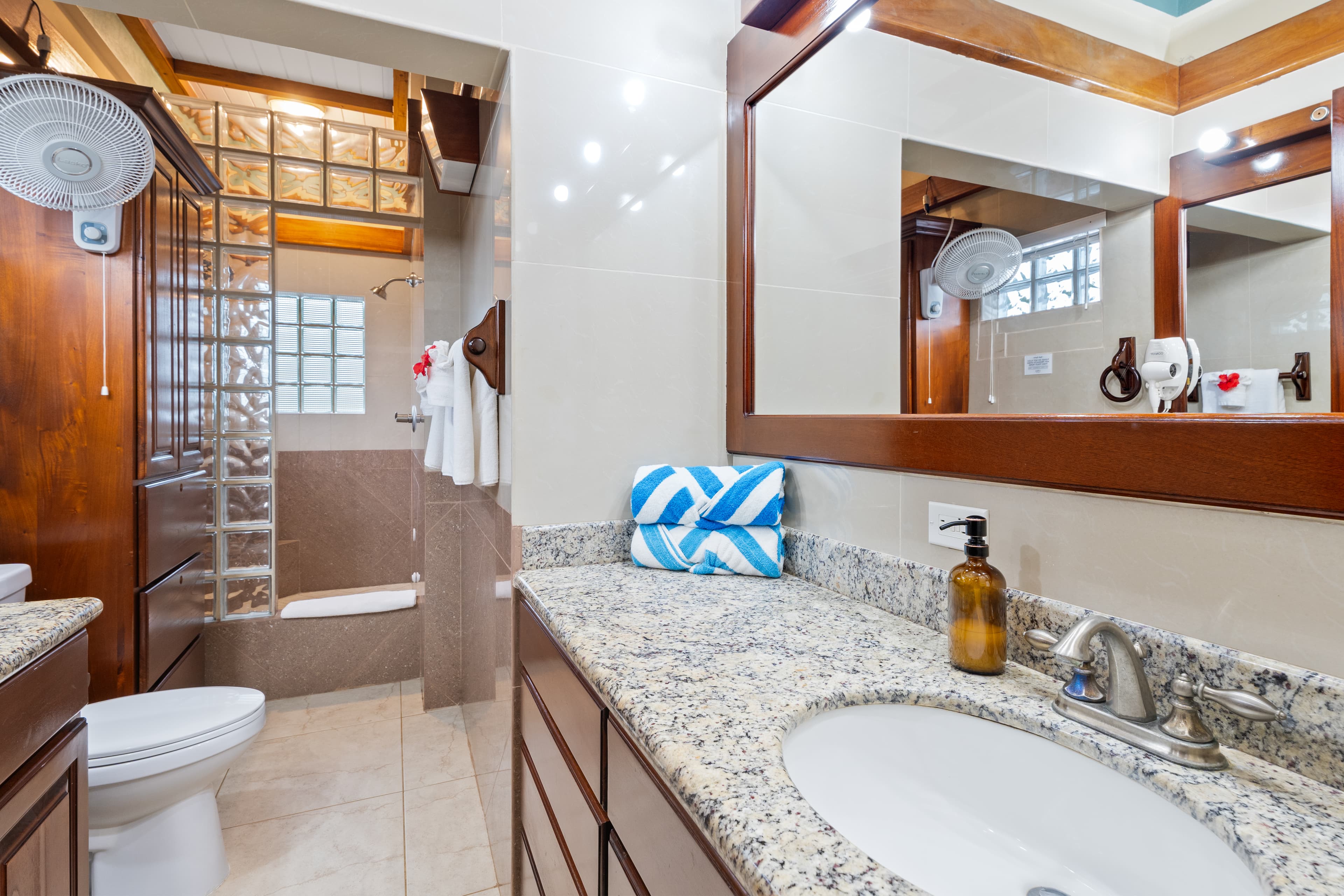 A modern bathroom with a granite vanity featuring a white undermount sink, a large wood-framed mirror, and blue-and-white patterned towels. In the background, a walk-in shower is visible behind a glass block wall, alongside a white toilet and dark wood cabinetry.