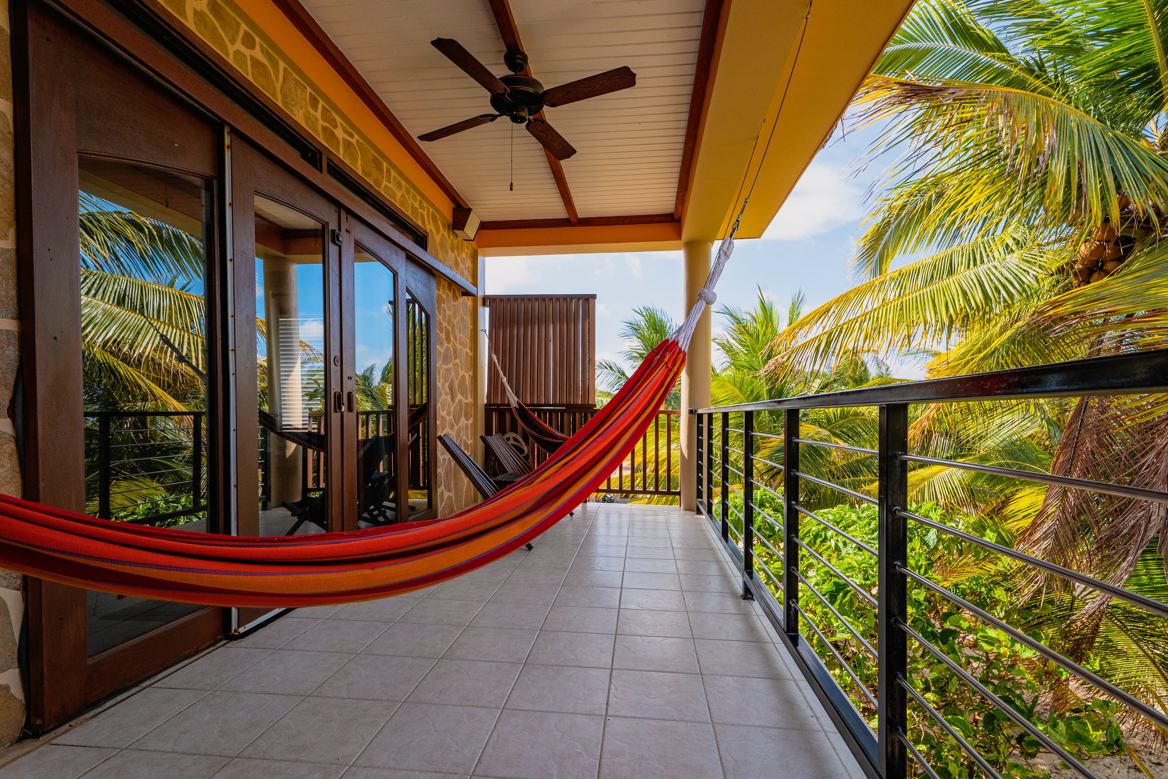 An outdoor balcony with tiled flooring and a dark wood-beamed ceiling featuring a ceiling fan. A red and orange striped hammock is stretched across the space, overlooking lush green palm trees under a bright sky. Dark wood-framed glass doors lead back into the room.