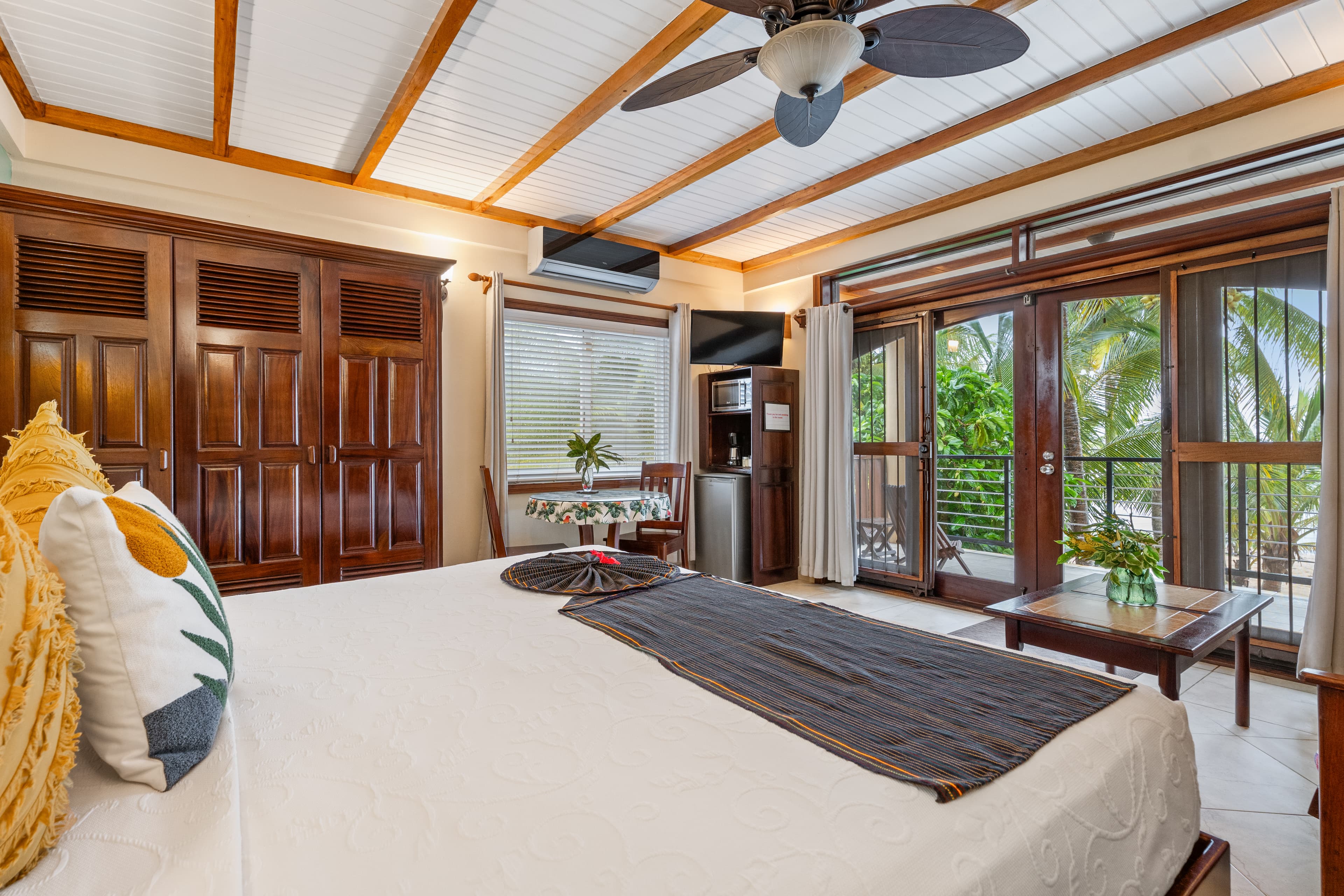A bedroom featuring a vaulted ceiling with exposed wooden beams and a white ceiling fan. The room includes a king bed with white linens and patterned pillows, rich dark wood cabinetry, and a sliding glass door that opens to a lush balcony with palm trees.