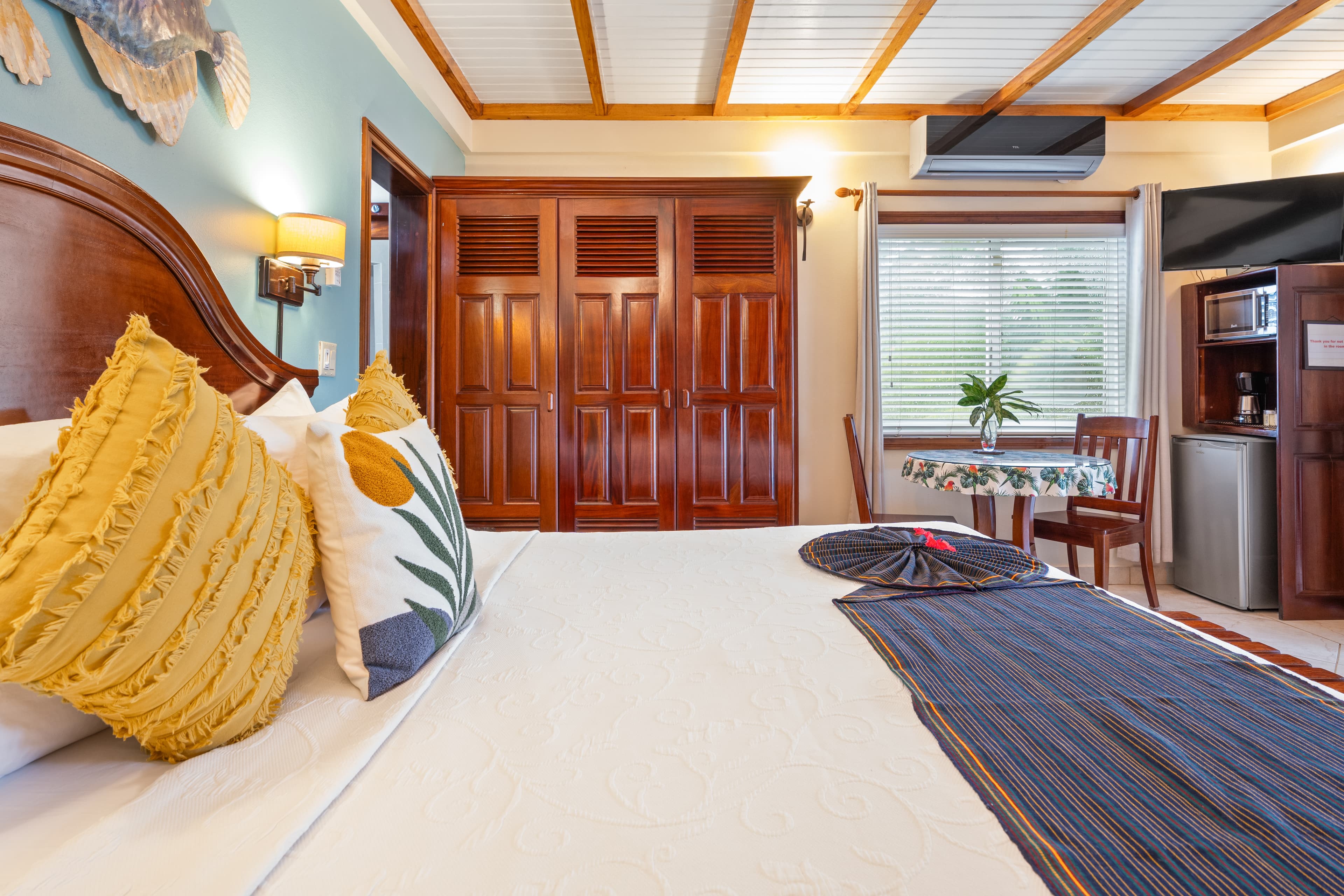 A tropical bedroom with a king bed dressed in white linens and blue-and-yellow pillows. The room features a light blue accent wall decorated with a large wooden fish carving and two small blue frog sculptures. A wooden bench sits at the foot of the bed, and a solid wood door is visible on the left.