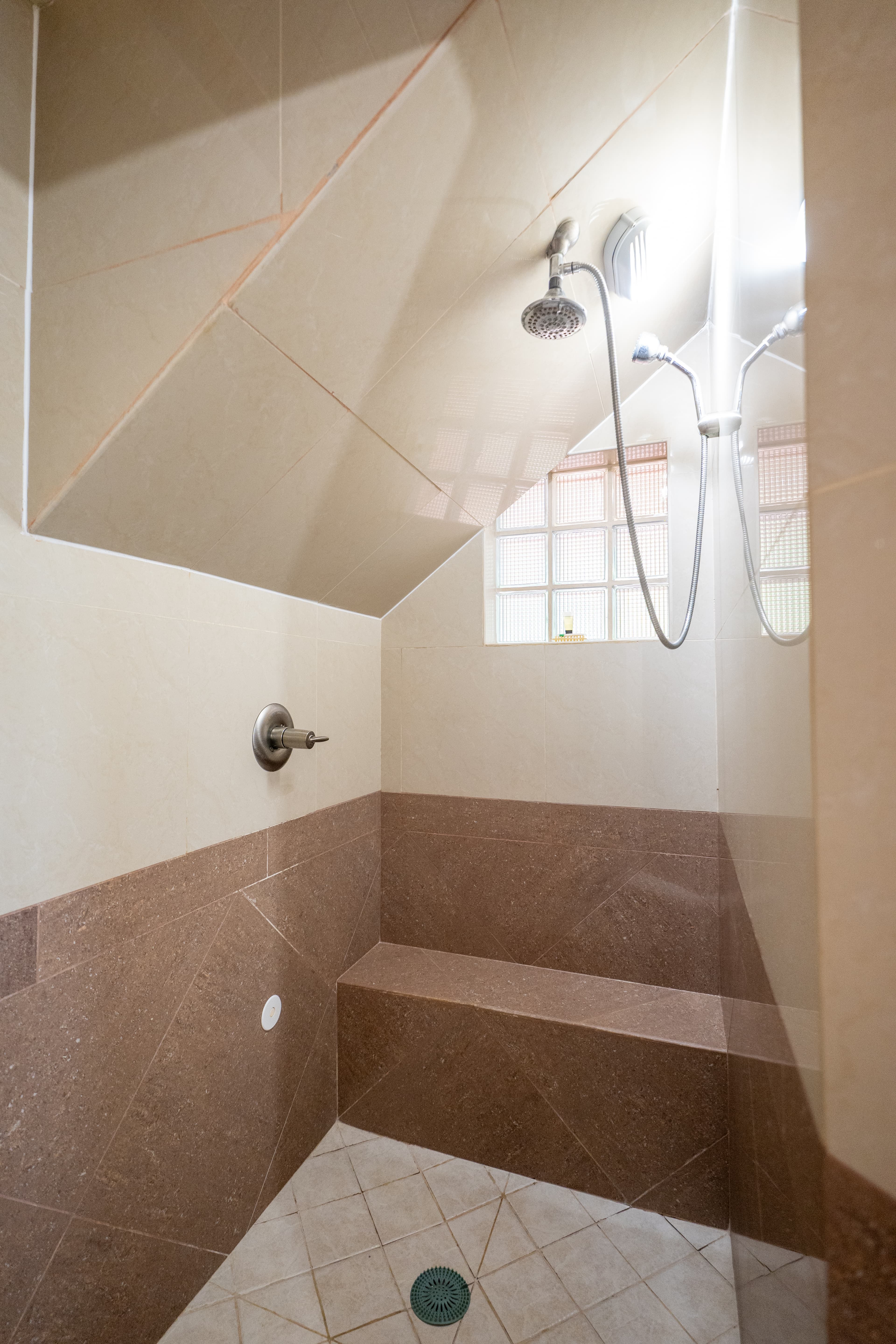 A modern walk-in shower featuring neutral-toned wall tiles and a built-in stone bench. The space includes a handheld showerhead, a glass block window for natural light, and a sloped ceiling following the architectural lines of the room.