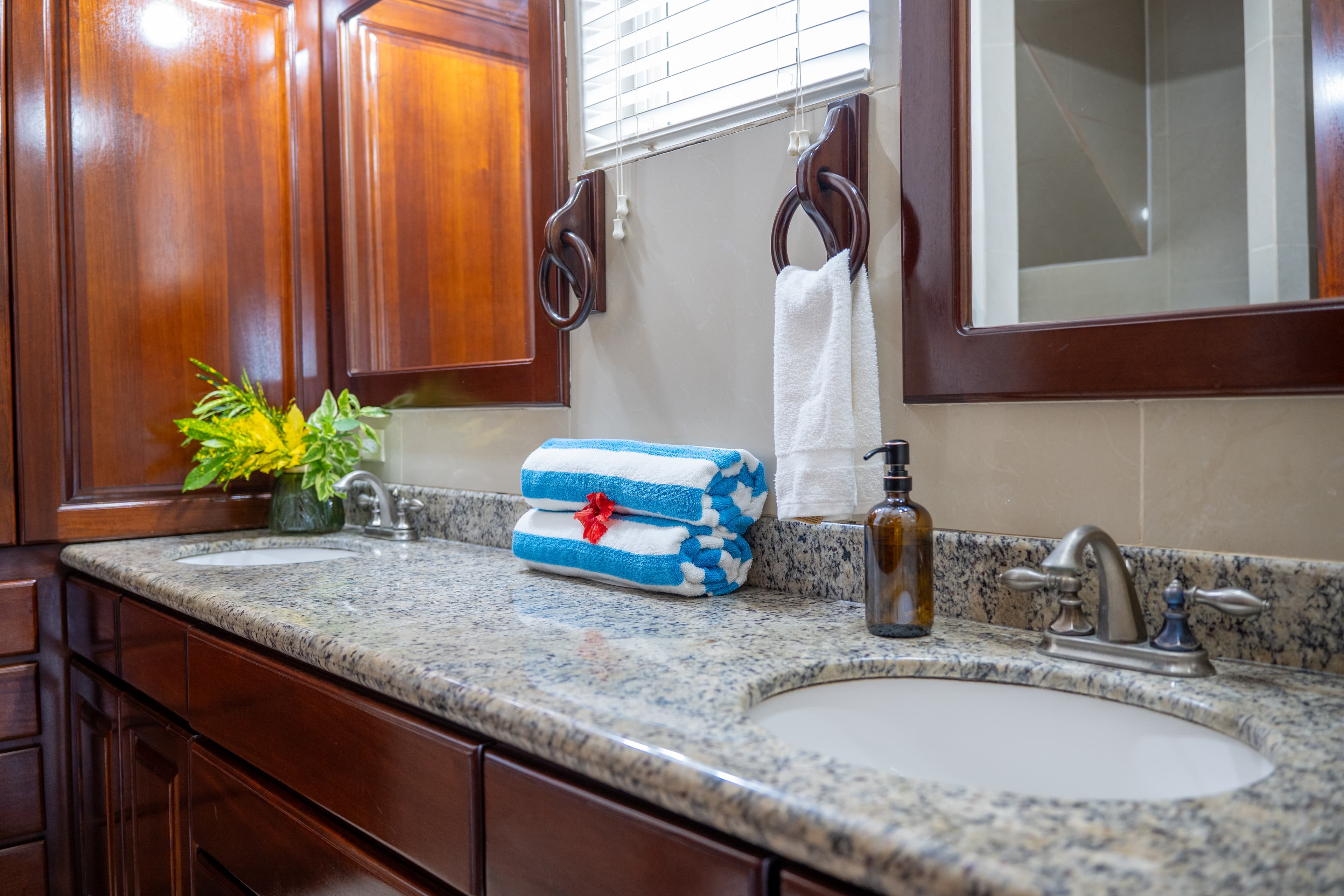 A double-sink bathroom vanity with a granite countertop and dark wood cabinetry. The counter is decorated with a vase of green and yellow tropical flowers, rolled blue-and-white striped towels with a red hibiscus flower, and a brown glass soap dispenser. Two dark wood-framed mirrors and a white towel hanging from a wooden ring are visible above the counter.