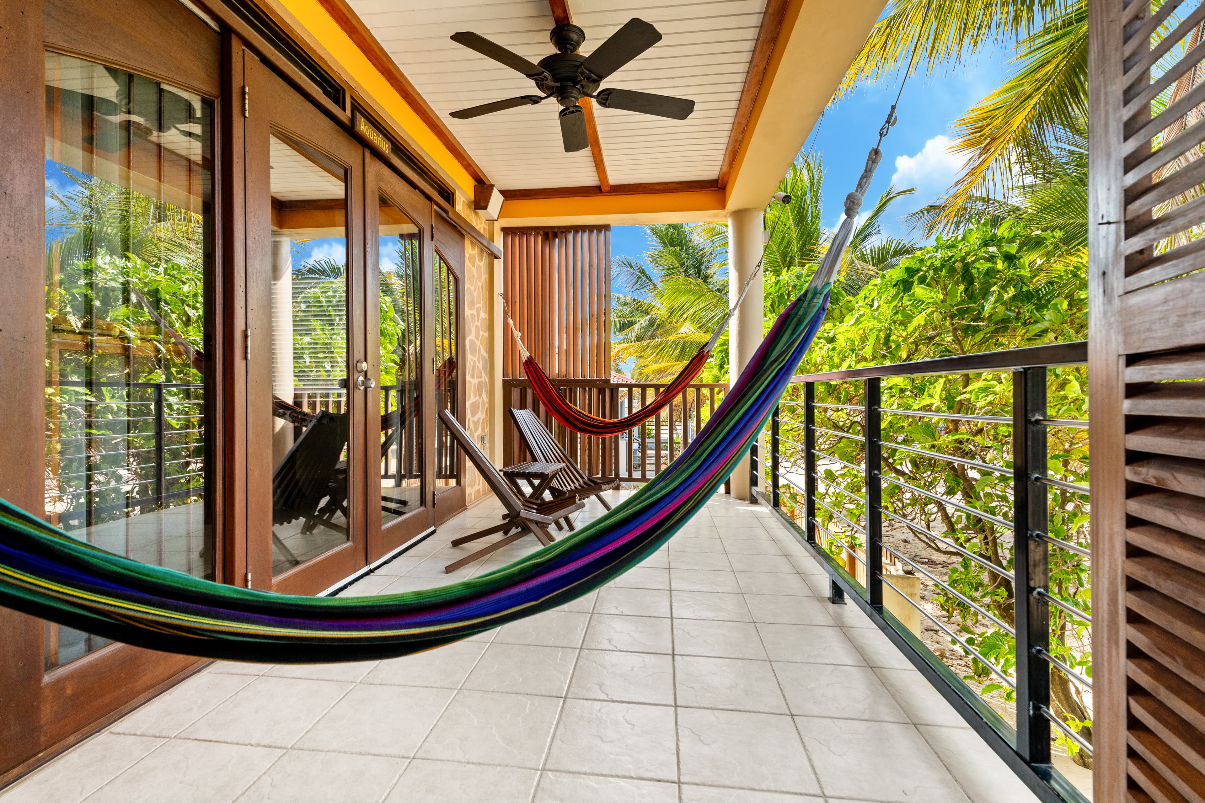 A spacious balcony with white tile flooring and a white ceiling featuring a dark wood ceiling fan. A colorful striped hammock is strung across the foreground, with two additional hammocks and wooden lounge chairs visible further down the deck. Large glass sliding doors with wooden frames lead inside, while a black metal railing overlooks lush green tropical foliage and palm trees under a blue sky.