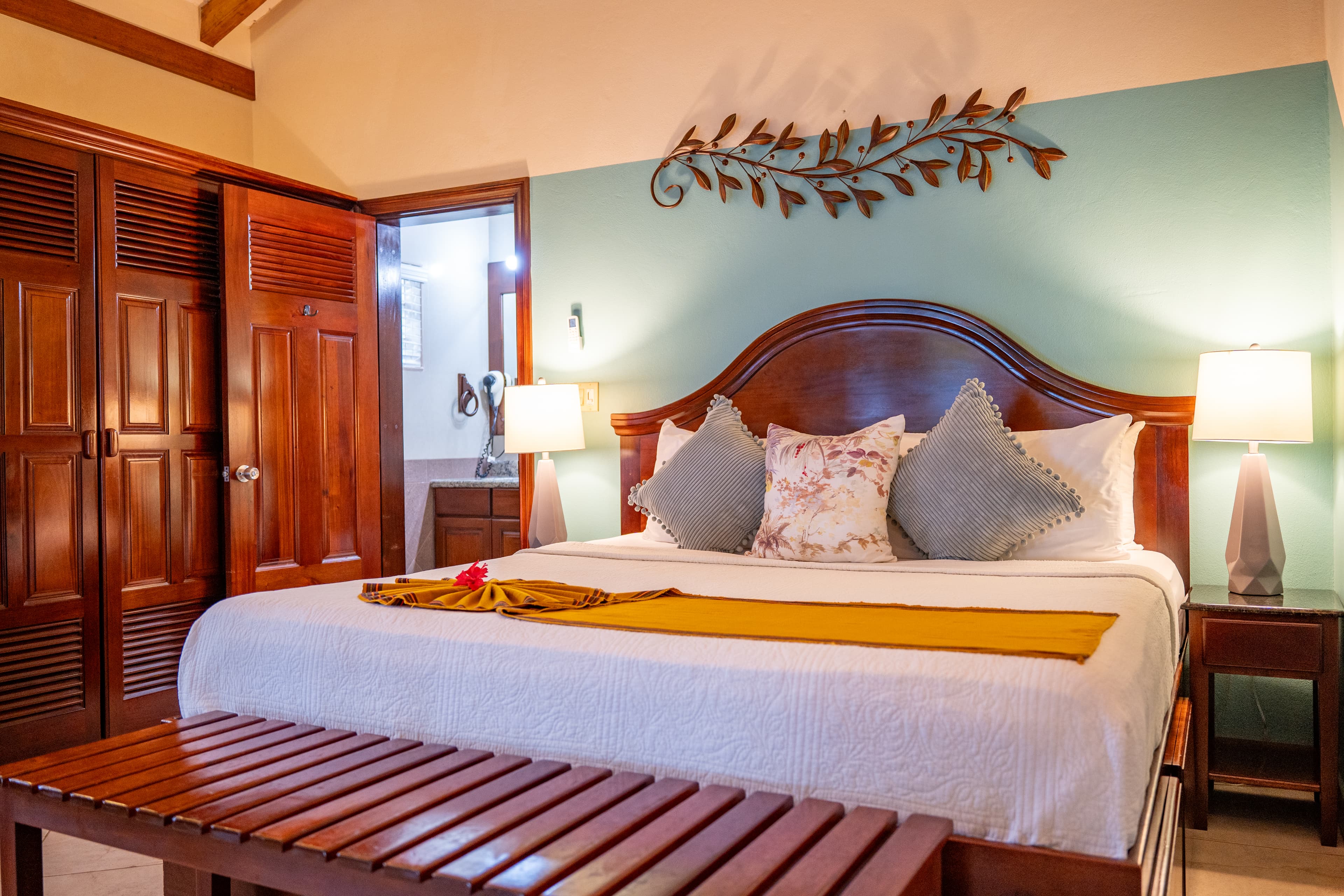 A tropical bedroom with a king bed dressed in white linens, featuring grey and floral accent pillows and a yellow runner. The room has a light blue accent wall adorned with a metal leaf-shaped wall hanging and is flanked by two bedside lamps. A wooden slatted bench sits at the foot of the bed, and a solid wood door and matching closet are visible on the left.