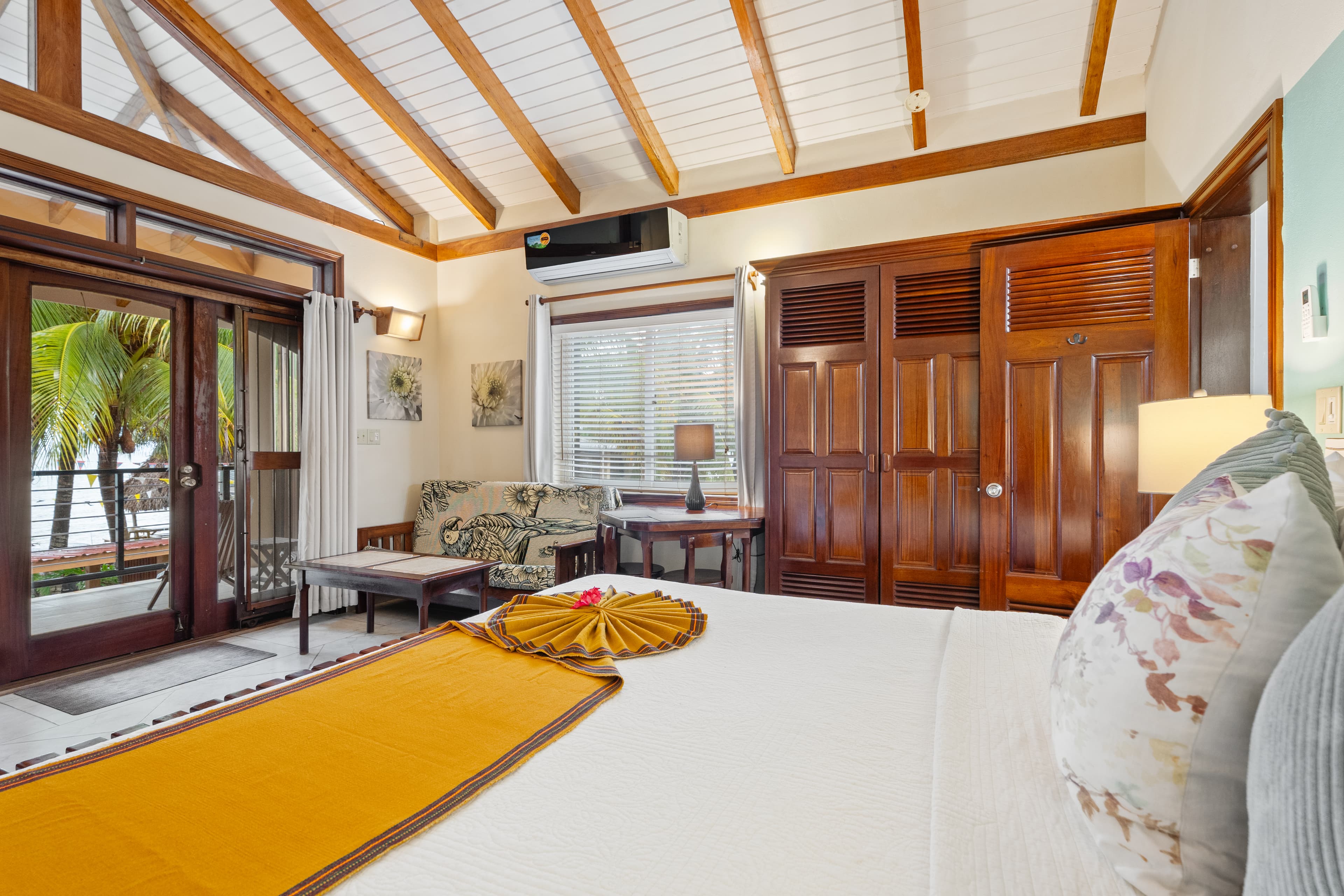 A tropical bedroom with a high vaulted ceiling featuring white panels and exposed wooden beams. The room contains a king bed with white linens, a bright yellow bed runner, and floral accent pillows. The interior is furnished with rich dark wood cabinetry, a small floral-patterned sofa, and a sliding glass door that leads to a balcony with palm trees.