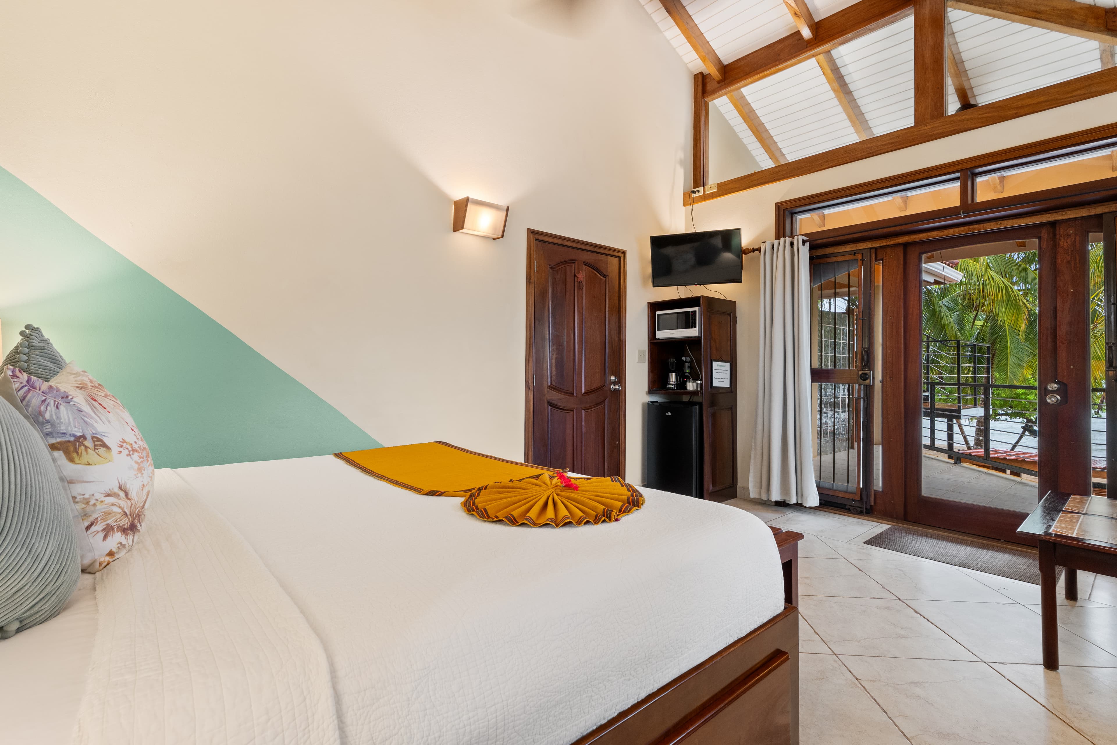 A tropical bedroom with a king bed featuring white linens, a bright yellow bed runner, and patterned accent pillows. The room has a vaulted ceiling with exposed wooden beams and is furnished with a dark wood door, a small kitchenette area with a microwave and mini-fridge, and a sliding glass door that opens to a balcony overlooking palm trees.