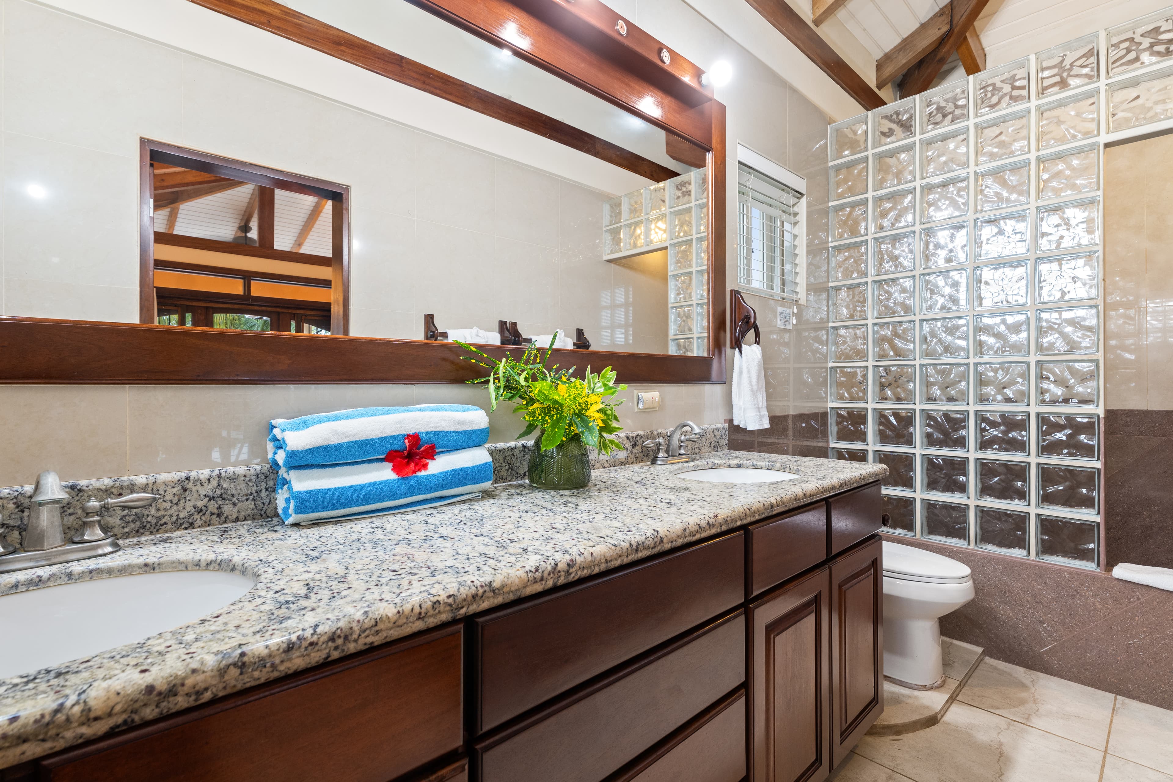A double-sink bathroom vanity with a granite countertop and dark wood cabinets, decorated with a vase of tropical flowers and rolled blue-and-white striped towels accented by a red hibiscus. The room features a large wood-framed mirror reflecting the vaulted ceiling and a decorative glass block wall next to the toilet.