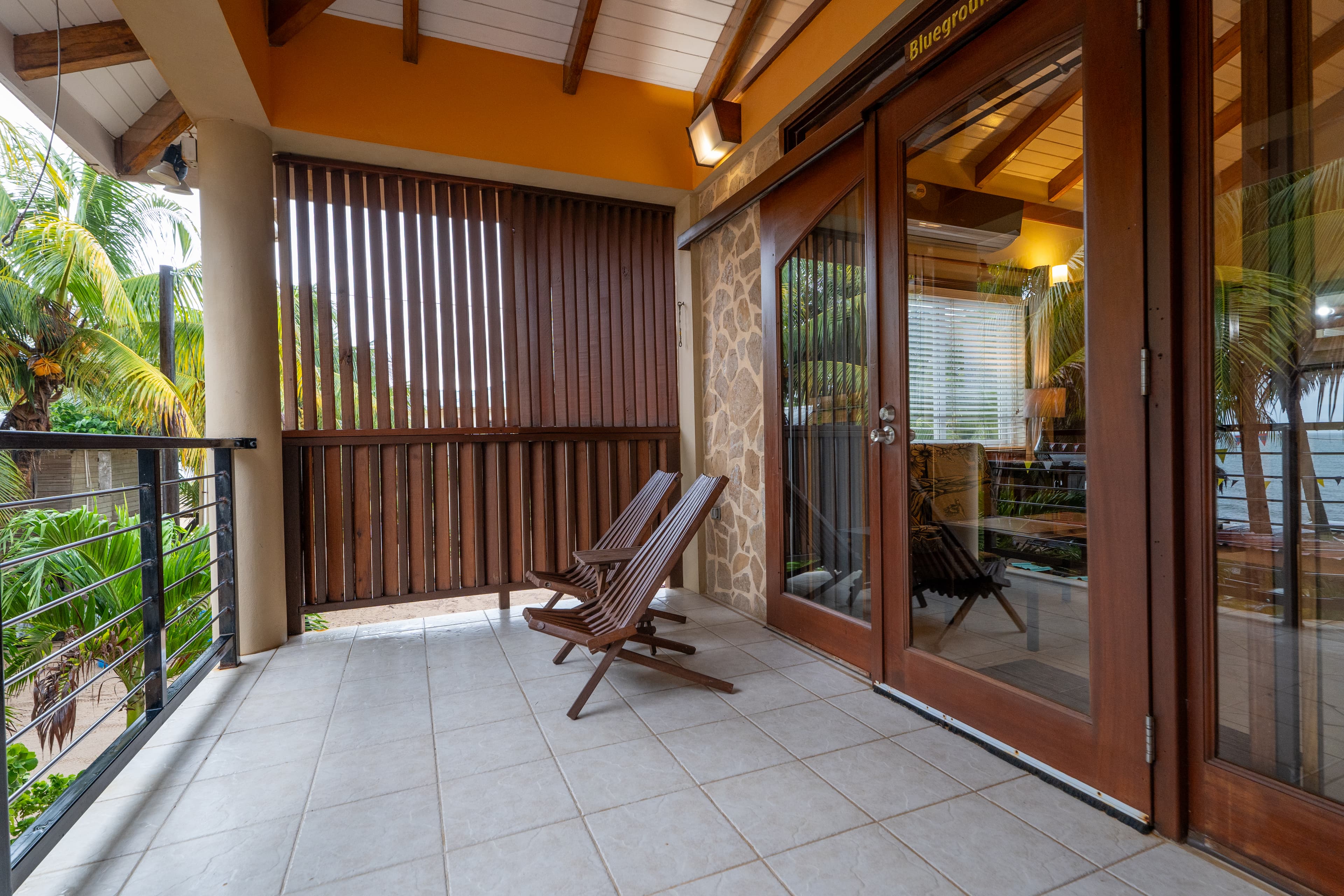 A balcony with white floor tiles and a wooden railing overlooking tropical palm trees. Two wooden lounge chairs sit on the deck next to large, wood-framed sliding glass doors that lead into a warmly lit interior.