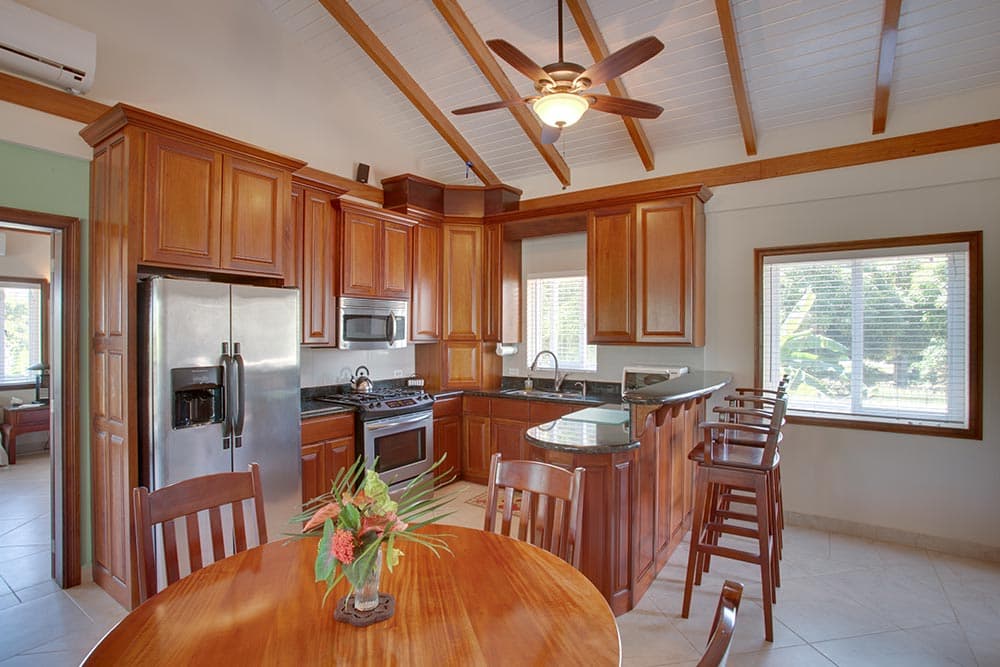 A tropical-style kitchen and dining area featuring rich wood cabinetry and a stainless steel refrigerator and oven. A round wooden dining table with a floral centerpiece sits in the foreground, while a breakfast bar with high-backed wooden chairs is positioned near a large window. The space is topped with a high vaulted ceiling with exposed wooden beams and a ceiling fan.