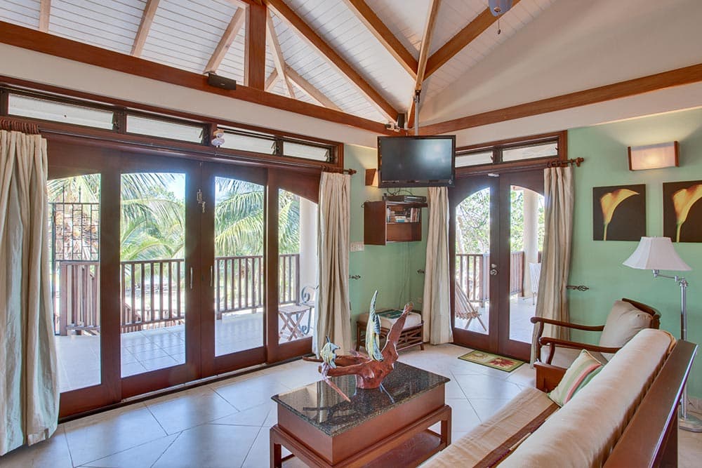 A tropical-style living area with a high vaulted ceiling featuring exposed wooden beams and a ceiling fan. Two sets of large, arched dark wood sliding glass doors open onto a spacious balcony overlooking lush palm trees. The room is furnished with a wooden sofa covered in a light neutral slipcover, a dark wood coffee table with a glass top, and light green accent walls decorated with framed botanical art.