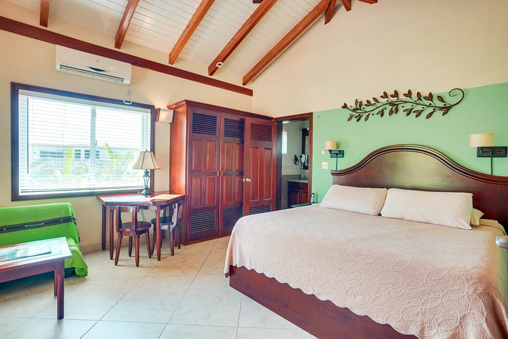 A tropical bedroom featuring a vaulted wood-beam ceiling with a ceiling fan, and a light green accent wall adorned with a decorative metal leaf wall hanging.