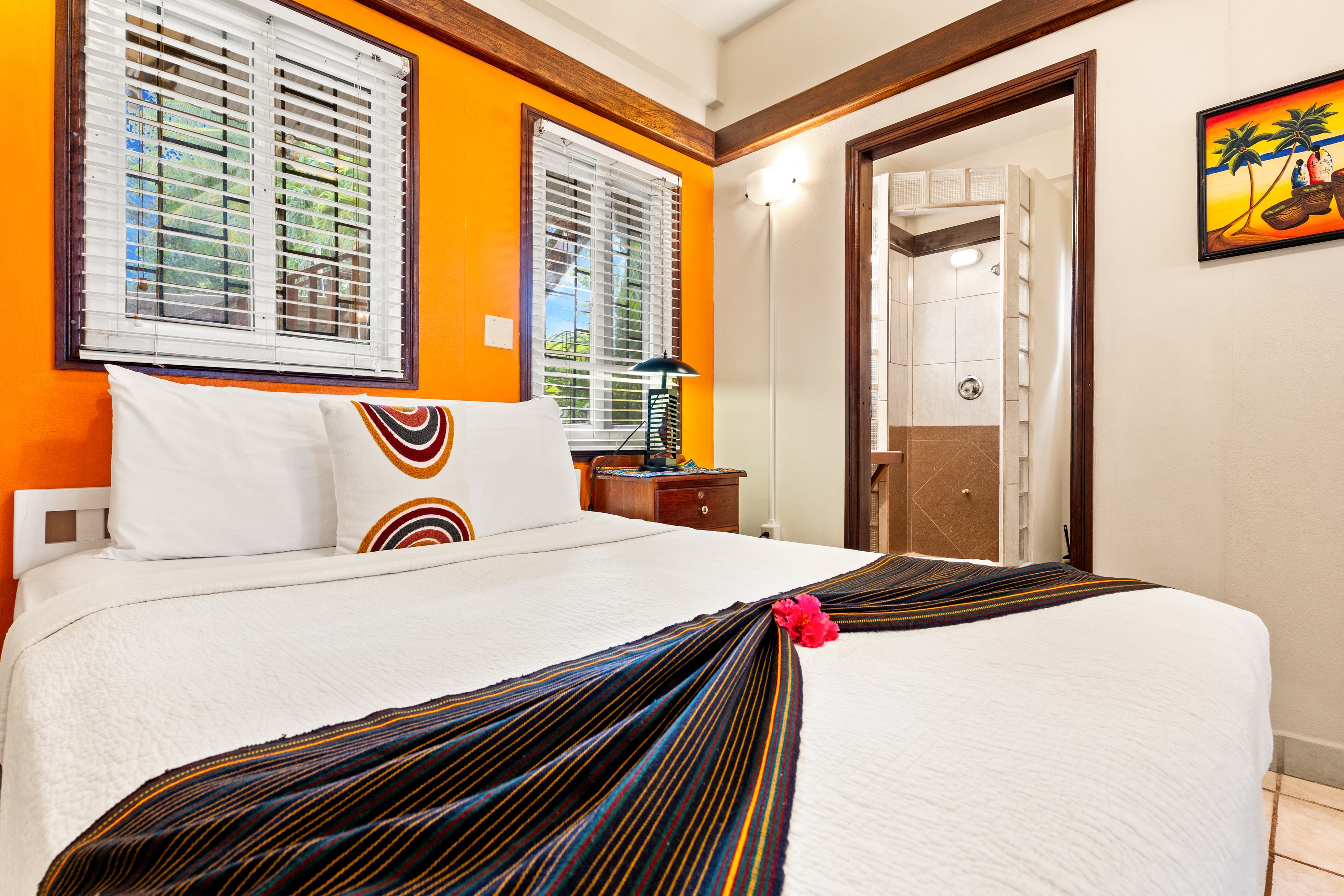 A cozy bedroom featuring bright orange accent walls and white linens on the bed, accented by a dark striped runner and a single red flower. The room includes two windows with white blinds, a dark wood nightstand with a lamp, and an open doorway leading to a brightly lit bathroom with a glass block shower wall.