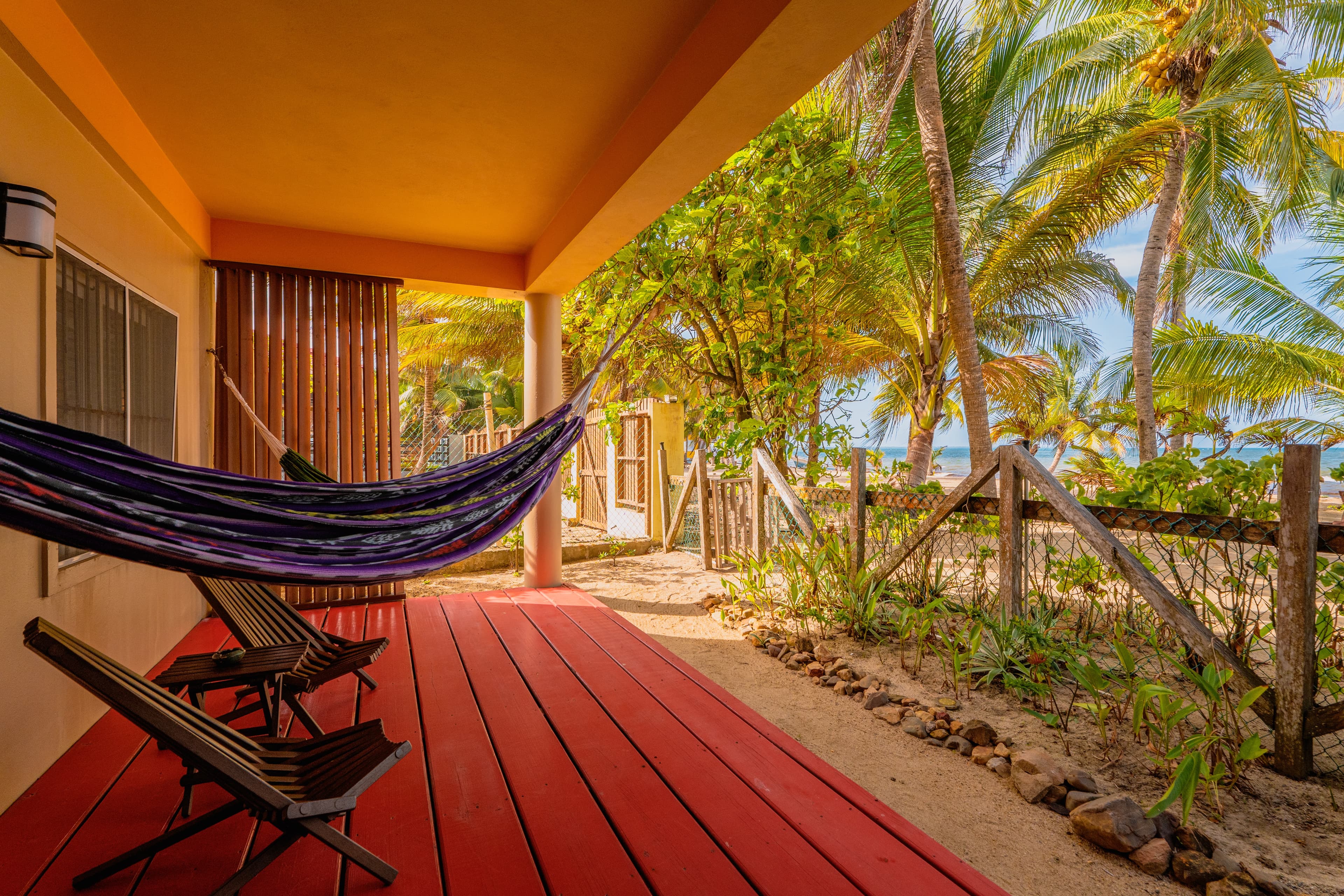 A shaded ground-floor patio with red decking, featuring a blue-and-white striped hammock and wooden lounge chairs. The patio overlooks a sandy area with lush tropical gardens and tall palm trees under a bright sky.