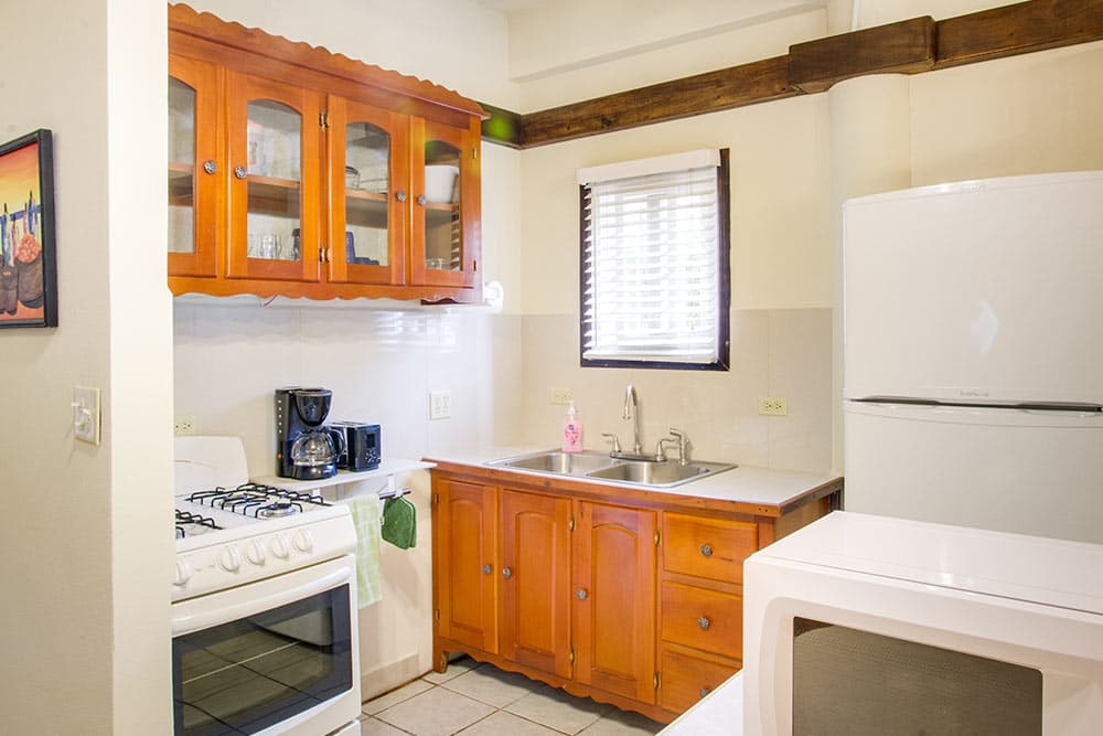 A small kitchen area with white appliances, including a stove, refrigerator, and microwave. The space features honey-toned wooden cabinetry with some glass-front doors, a double stainless steel sink under a window with white blinds, and a neutral tiled backsplash.