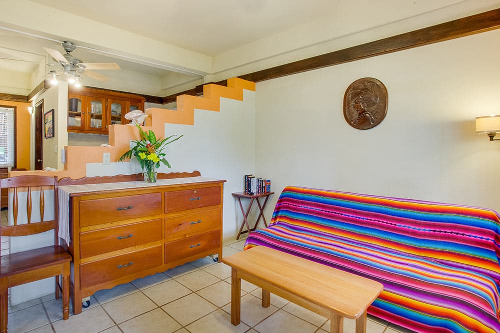 A tropical-themed living space with white tiled floors and multiple ceiling fans. The room features a wooden dresser topped with a fresh floral arrangement, a sofa covered in a colorful, horizontally striped blanket, and a white staircase with wooden treads in the background.