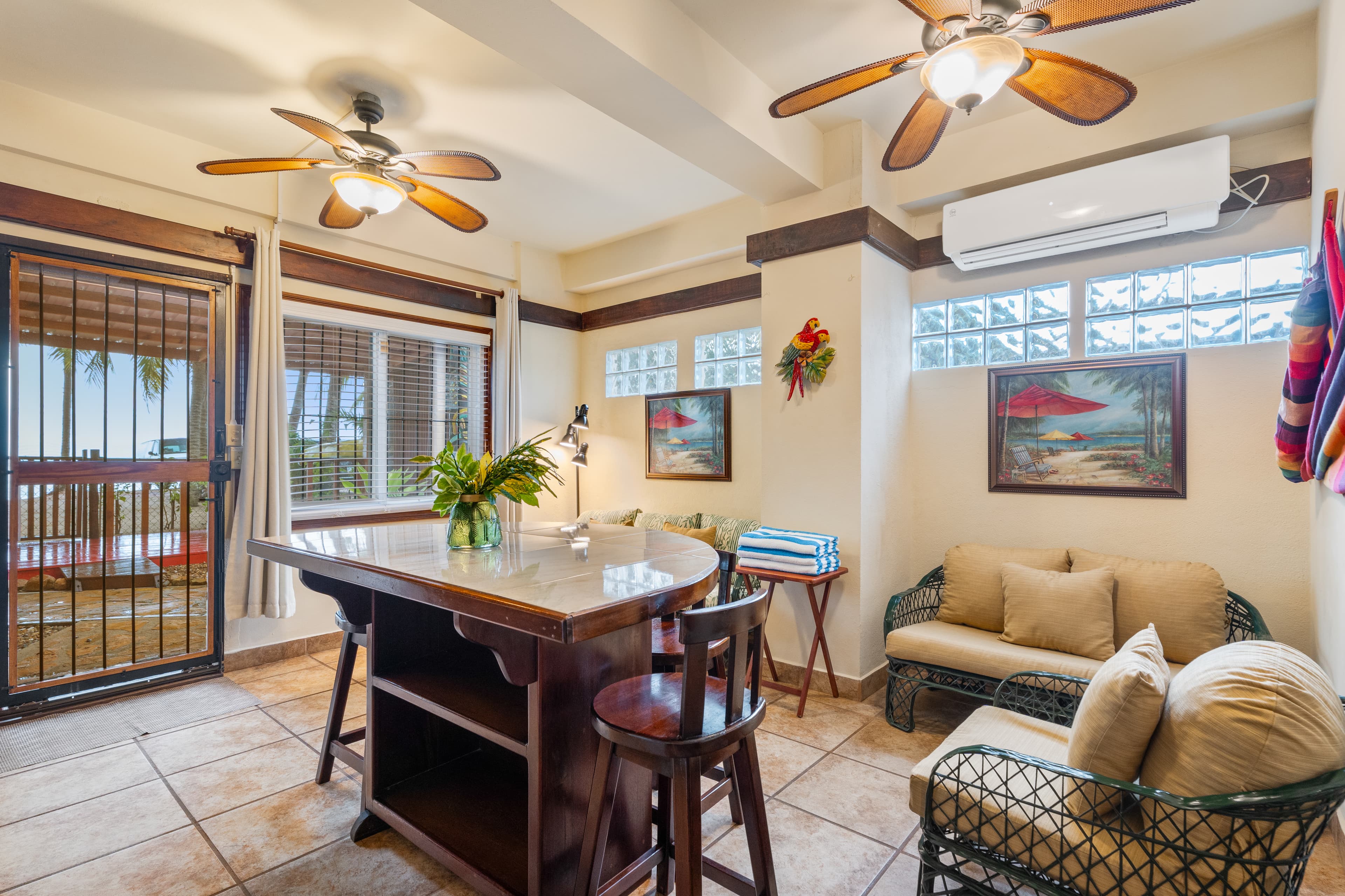 A tropical living area featuring white tiled floors, multiple ceiling fans, and neutral-colored walls. The space is furnished with a dark wood breakfast bar with stools, a small wicker sofa with tan cushions, and a second matching wicker armchair. Two framed paintings of beach scenes hang on the walls, and a glass door with a security screen leads to a sunlit balcony.
