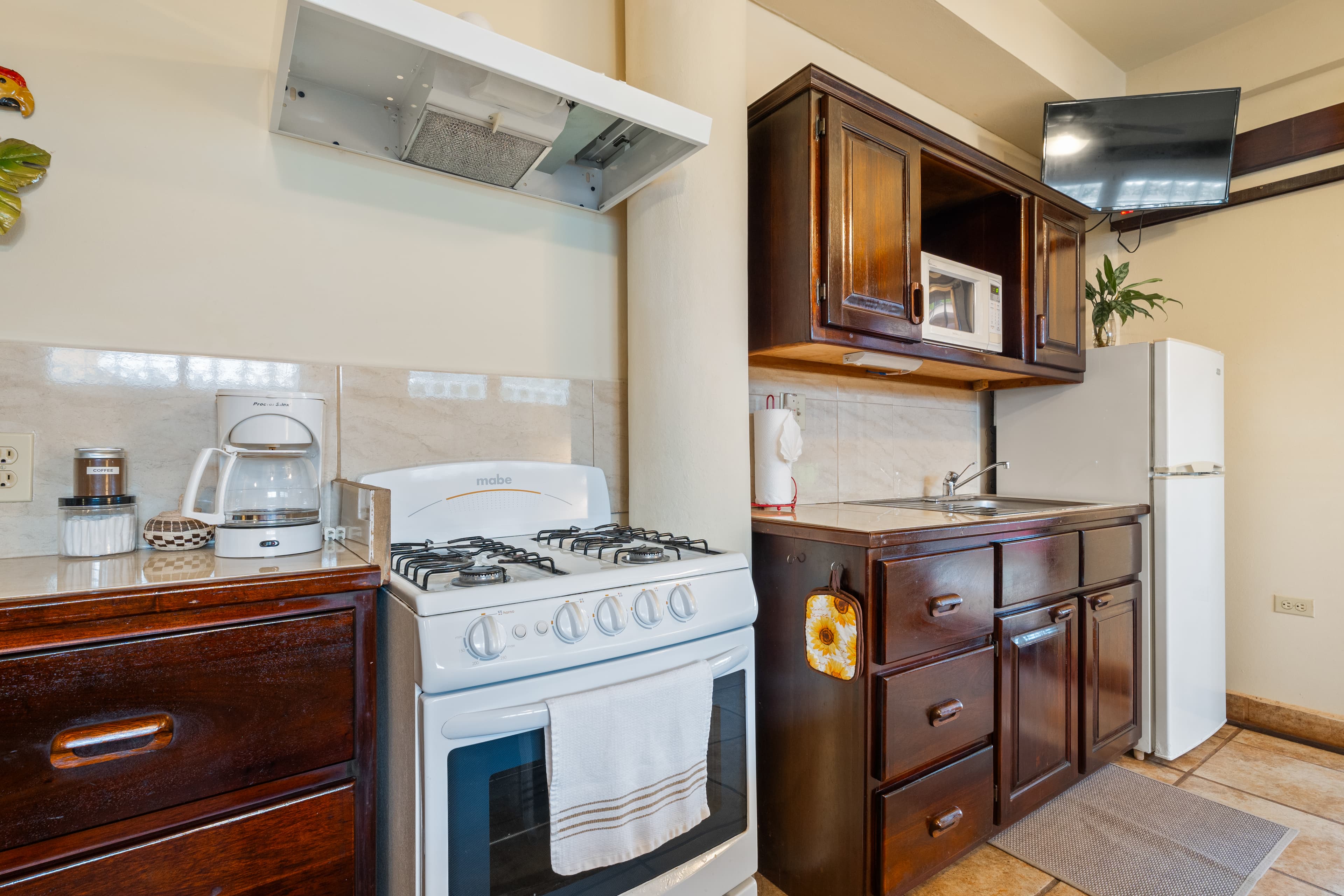 A compact kitchen with dark wood cabinetry and a white refrigerator and gas stove. The counter is equipped with a white coffee maker, a toaster, and a built-in microwave, with a flat-screen television mounted above the cabinets.
