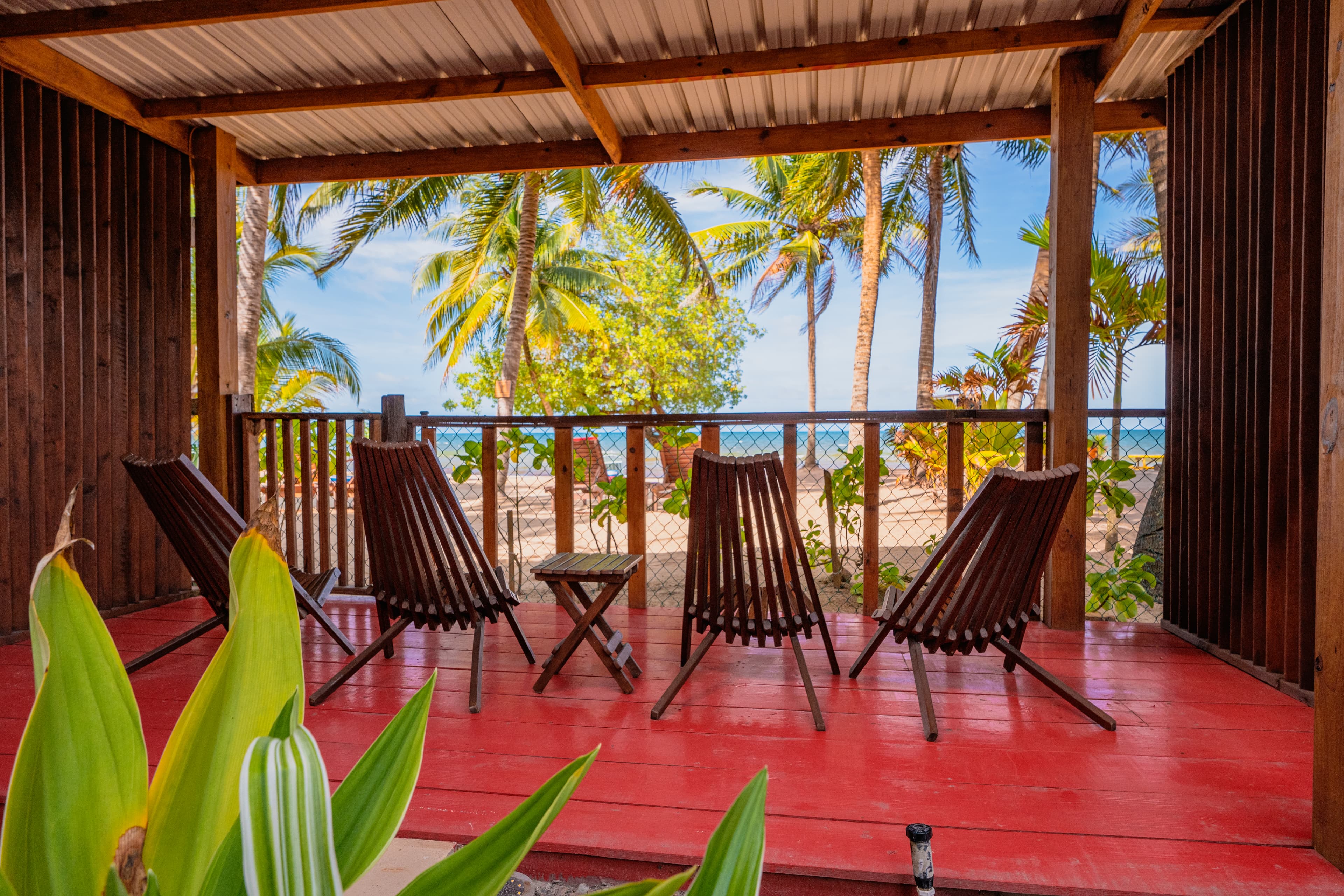 A shaded ground-floor patio with a vibrant red floor and dark wooden slatted lounge chairs. The patio is enclosed by a wooden railing and overlooks a sunny beach with tall palm trees and a view of the turquoise ocean in the distance.