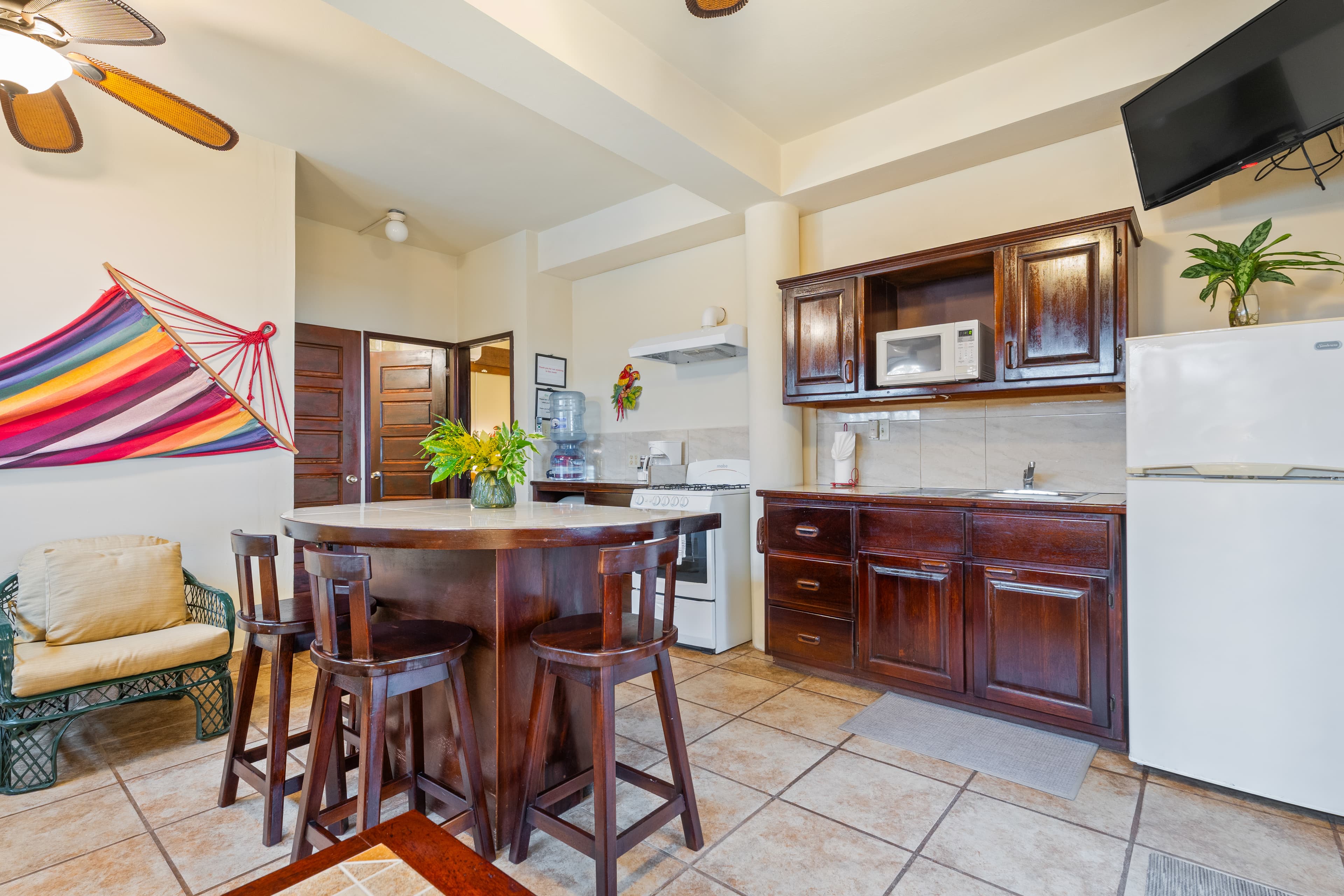 A tropical-themed bedroom featuring a wooden bed with white linens and yellow accent pillows, a large decorative fish on a light blue wall, and wooden ceiling beams with a fan. A wooden bench sits at the foot of the bed, and two blue frog decorations are mounted near a dark wood door.