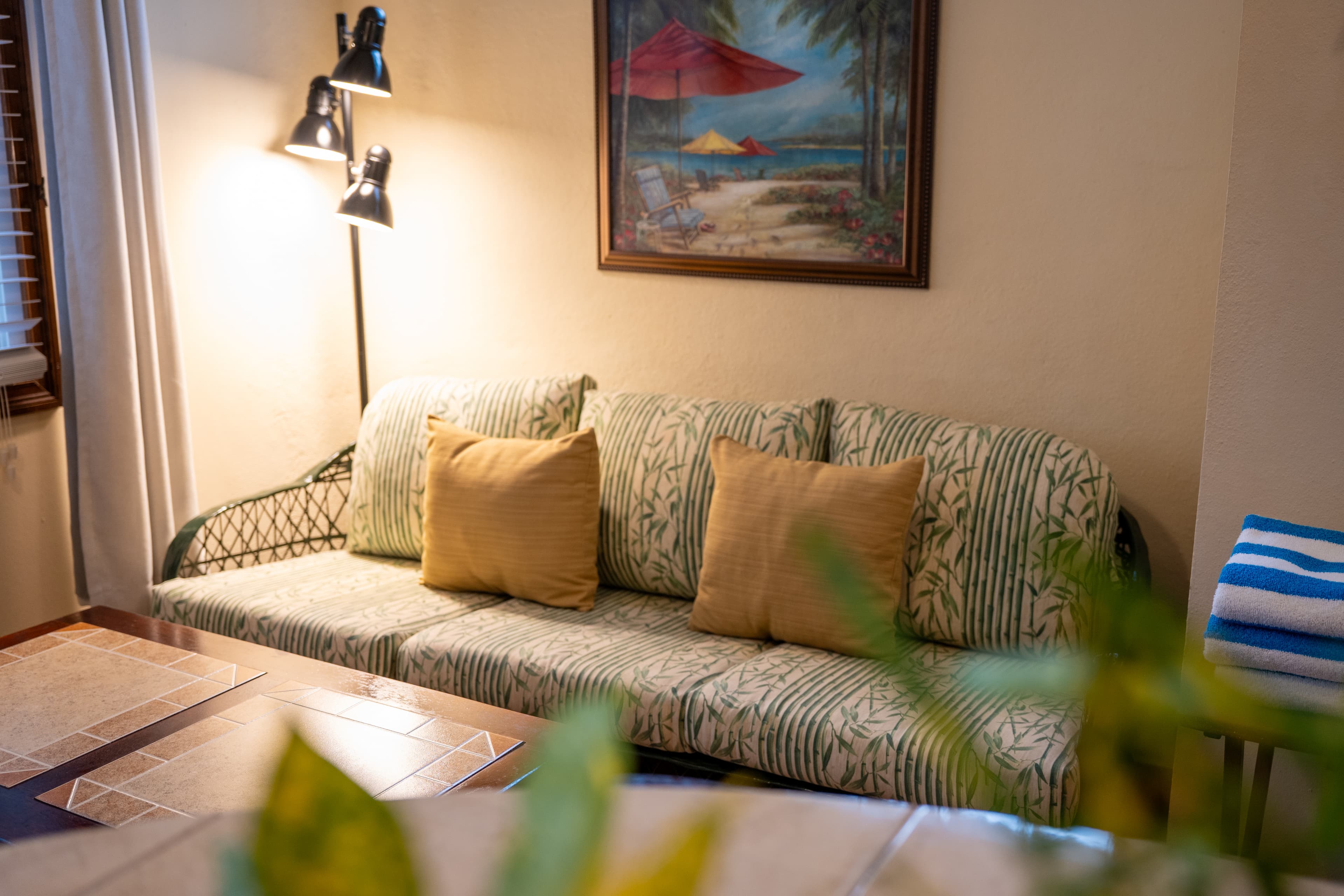 A shaded patio with vibrant red flooring, featuring four dark wooden slatted chairs and a small wooden side table. The space is enclosed by a dark wood railing, providing a direct view of a sandy beach lined with lush palm trees and the turquoise ocean under a bright, clear sky.