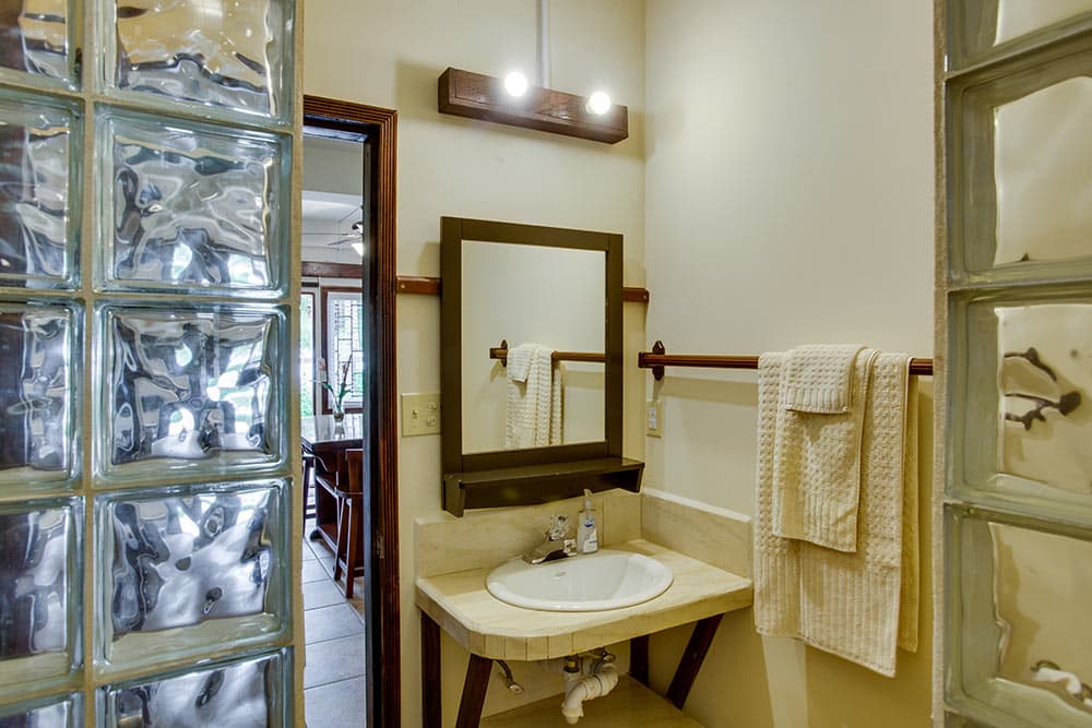 A tropical-themed bathroom featuring a cream-colored pedestal sink with a dark wood-framed mirror. The space includes a glass block partition wall for the shower and a window with frosted glass slats, with white towels hanging from a wooden rail.