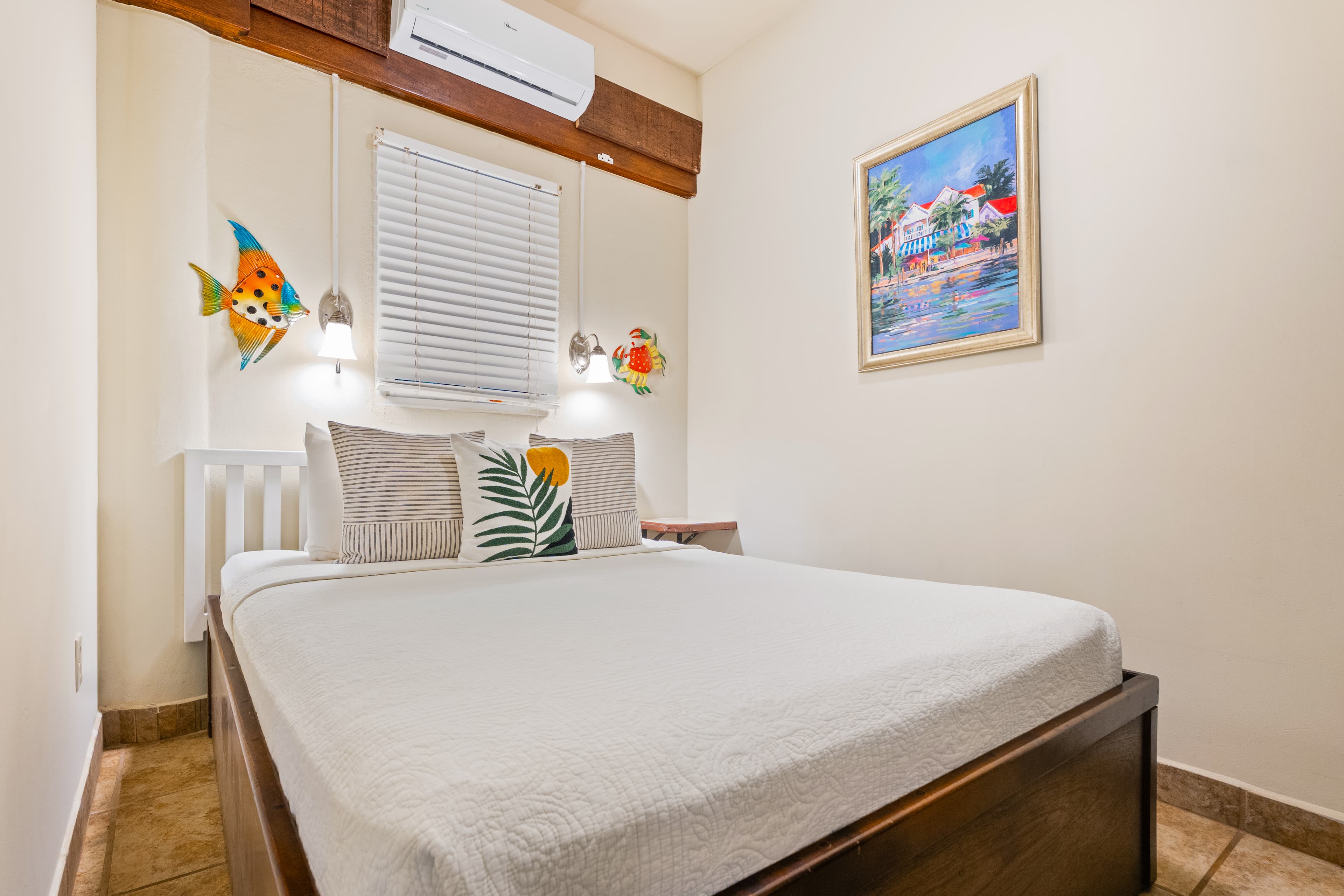 A tropical-style bedroom with white linens on a queen bed, featuring yellow accent pillows and a dark striped runner with a red hibiscus. The room is decorated with a large wooden fish carving and two blue frog sculptures on a light blue accent wall, balanced by dark wood cabinetry and a high vaulted ceiling with exposed beams.