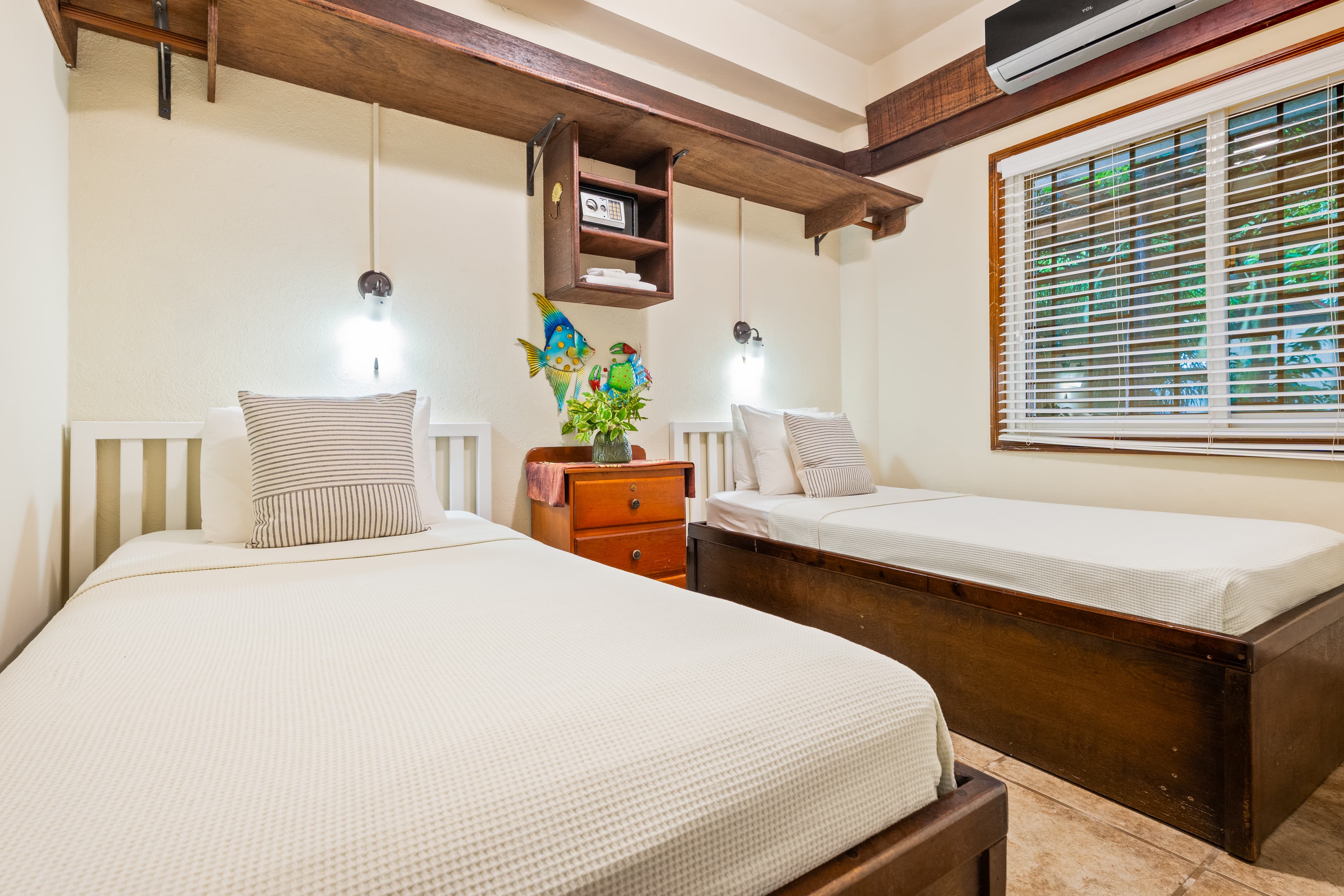 A tropical-style bedroom featuring two twin beds with white linens and grey accent pillows. The room has neutral walls with dark wood trim, a central wooden nightstand topped with a small fish decoration and flowers, and a large window with white blinds. Wall-mounted reading lamps are positioned above each bed, and a wooden shelf runs along the top of the back wall.