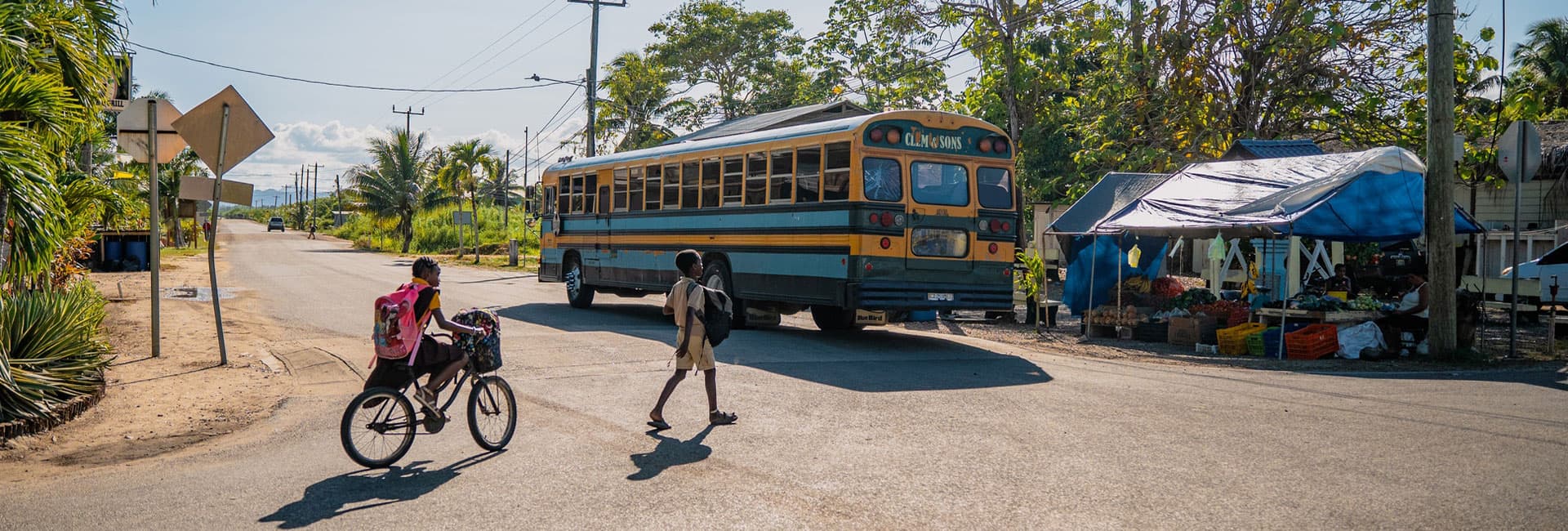 A cyclist and a pedestrian cross a road near a bus stop and outdoor market.