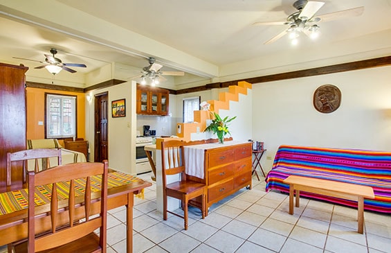 A vibrant living space featuring white tiled floors and multiple ceiling fans. The room is furnished with a wooden dining set with a striped runner, a wooden dresser with a fresh floral arrangement, and a sofa covered in a colorful, horizontally striped blanket. A white staircase with wooden treads is visible in the background, along with a small kitchen area and orange accent walls.