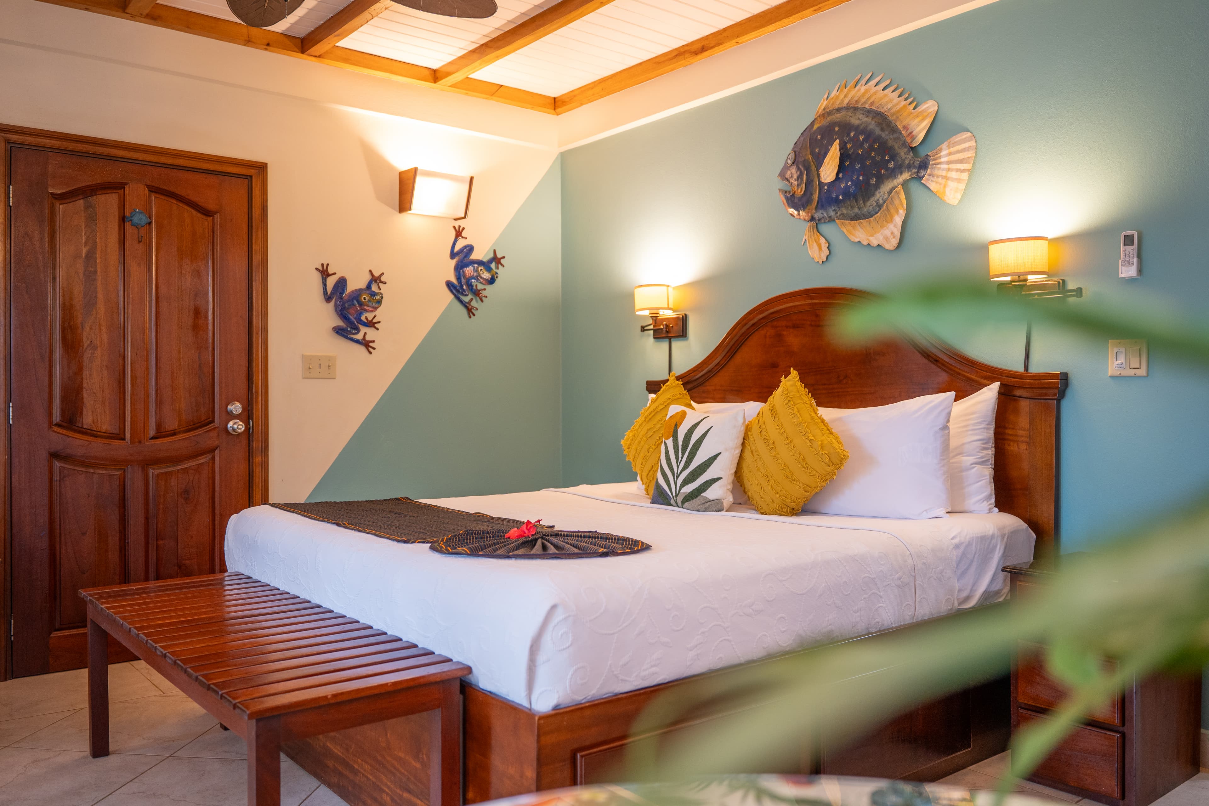 A tropical-themed bedroom featuring a wooden bed with white linens and yellow accent pillows, a large decorative fish on a light blue wall, and wooden ceiling beams with a fan. A wooden bench sits at the foot of the bed, and two blue frog decorations are mounted near a dark wood door.