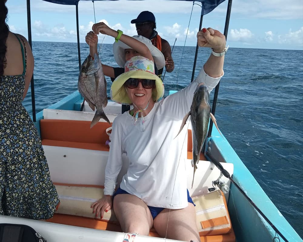 A woman wearing a sun hat proudly holds two freshly caught fish while sitting on a boat.