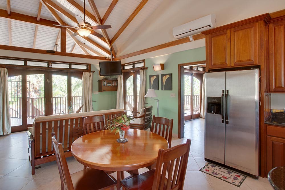 A tropical-themed living area with a high vaulted ceiling, exposed wooden beams, and a ceiling fan. The room features a wooden sofa with a neutral cover, a dark wood dining table with four chairs, and a stainless steel refrigerator in a kitchen area with wood cabinetry. Two sets of arched wooden sliding glass doors lead to a balcony with lush green views.