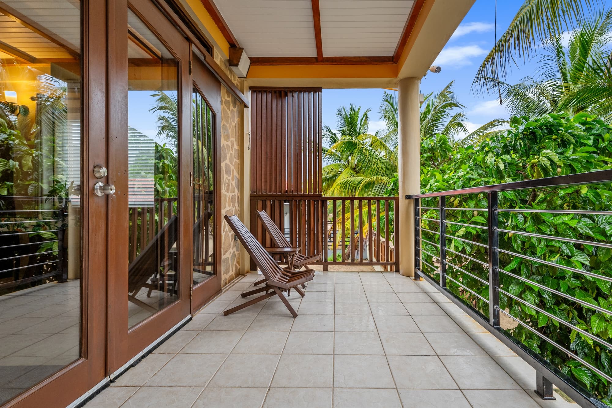A balcony with two wooden chairs overlooks lush greenery and palm trees.