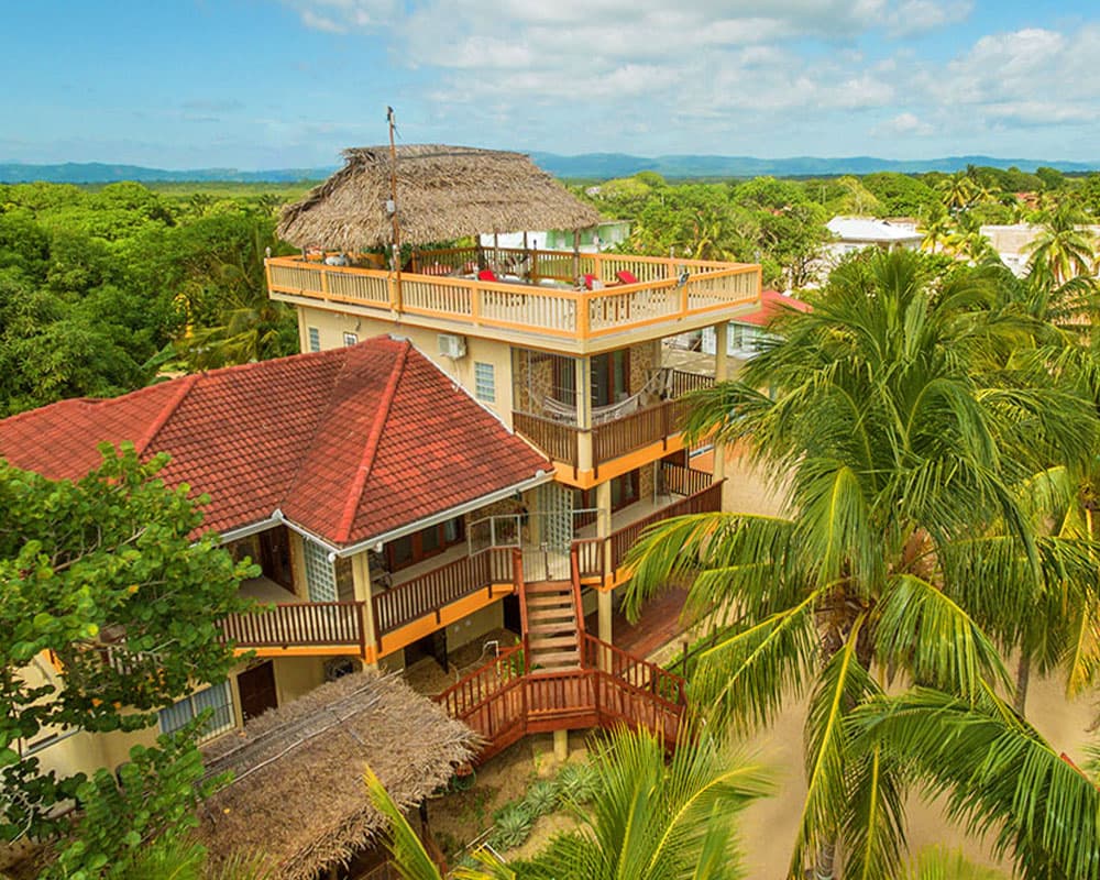A two-story house with a thatched roof surrounded by lush greenery and palm trees.