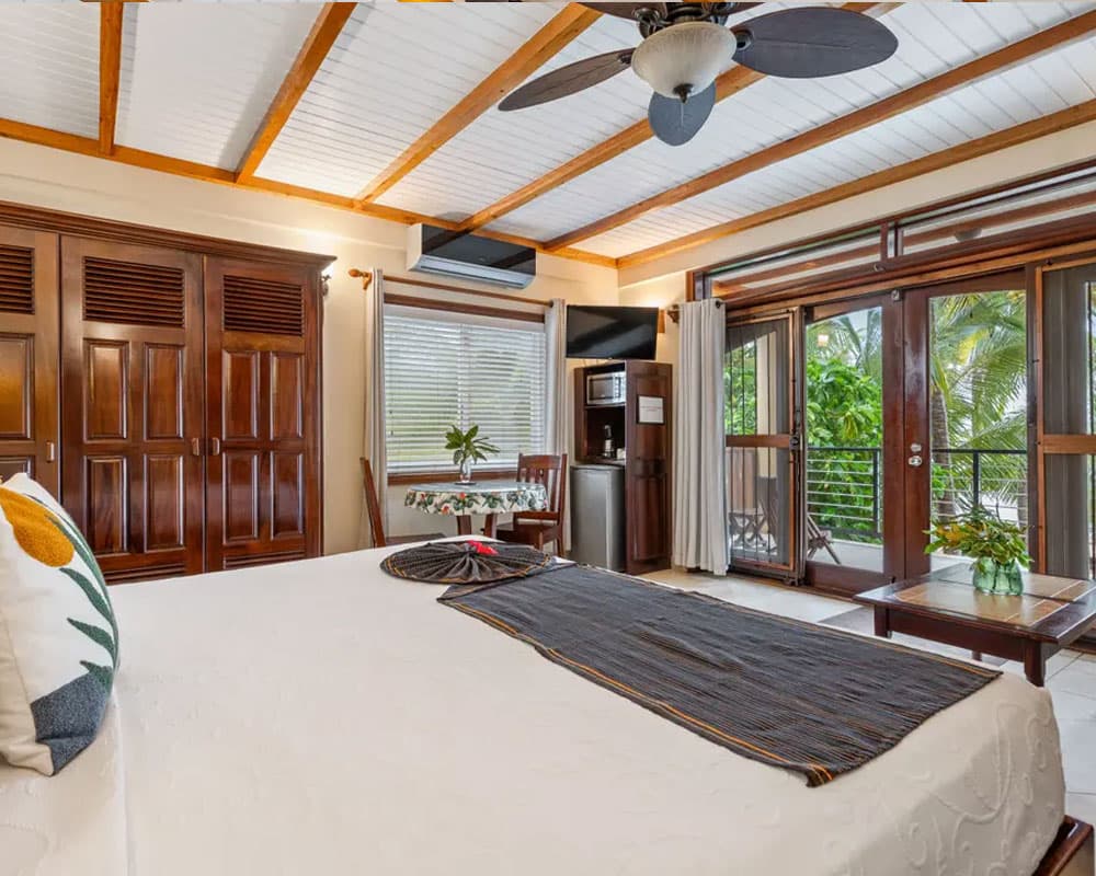 A cozy bedroom with wooden accents, featuring a king-size bed, a small fridge, and access to a balcony surrounded by greenery.