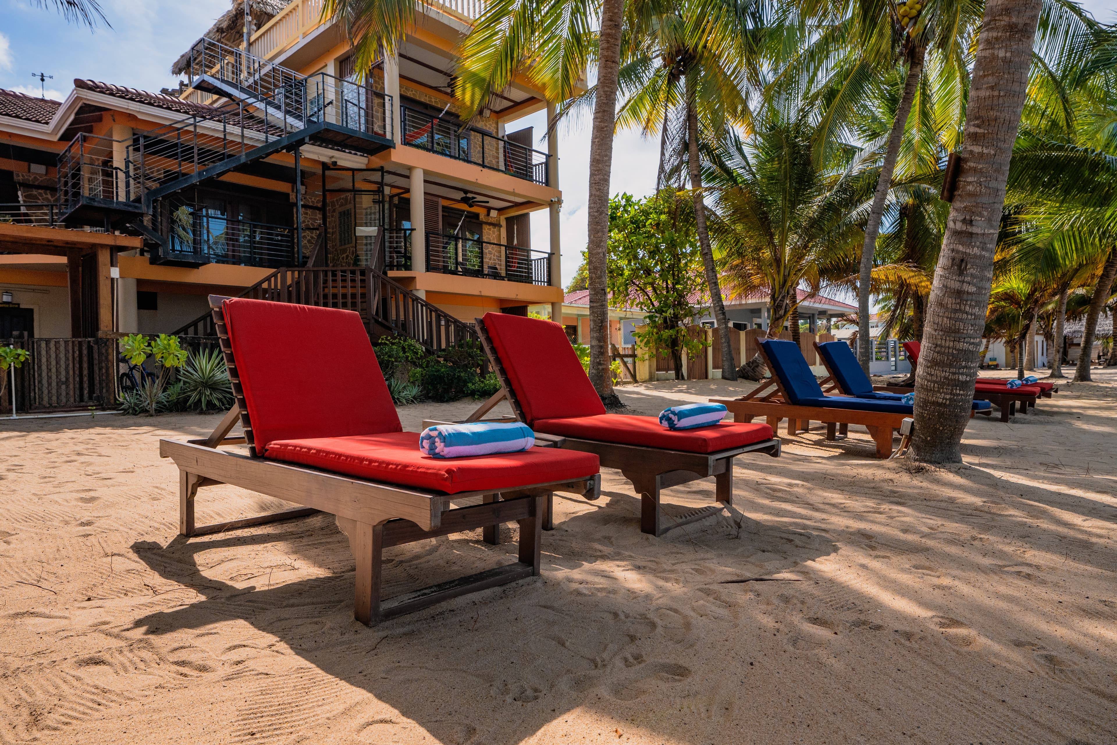 Lounge chairs with red cushions on a sandy beach in front of a multi-story building surrounded by palm trees.
